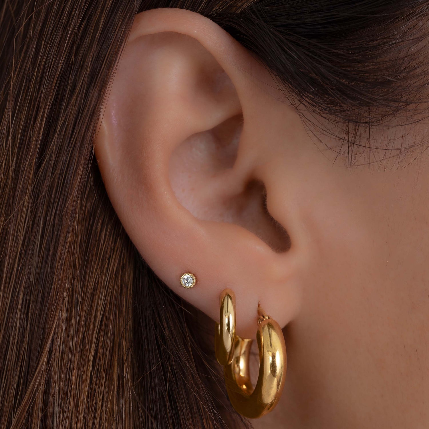Model wearing trendy gold thick tube hoop earrings paired with minimalist hoop earrings and a diamond bezel stud earring.