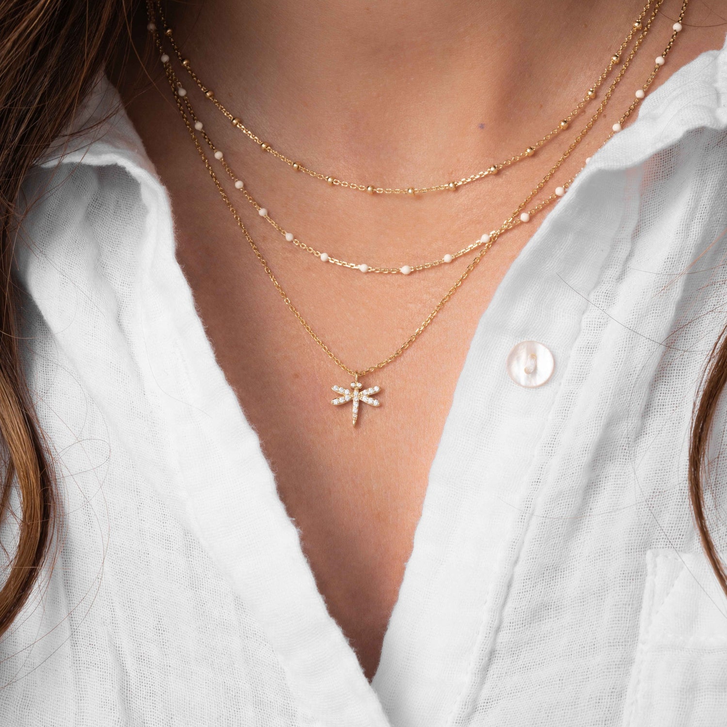 model wearing dainty dragonfly necklace paired with a cream enamel beaded necklace and a gold simple beaded chain necklace.