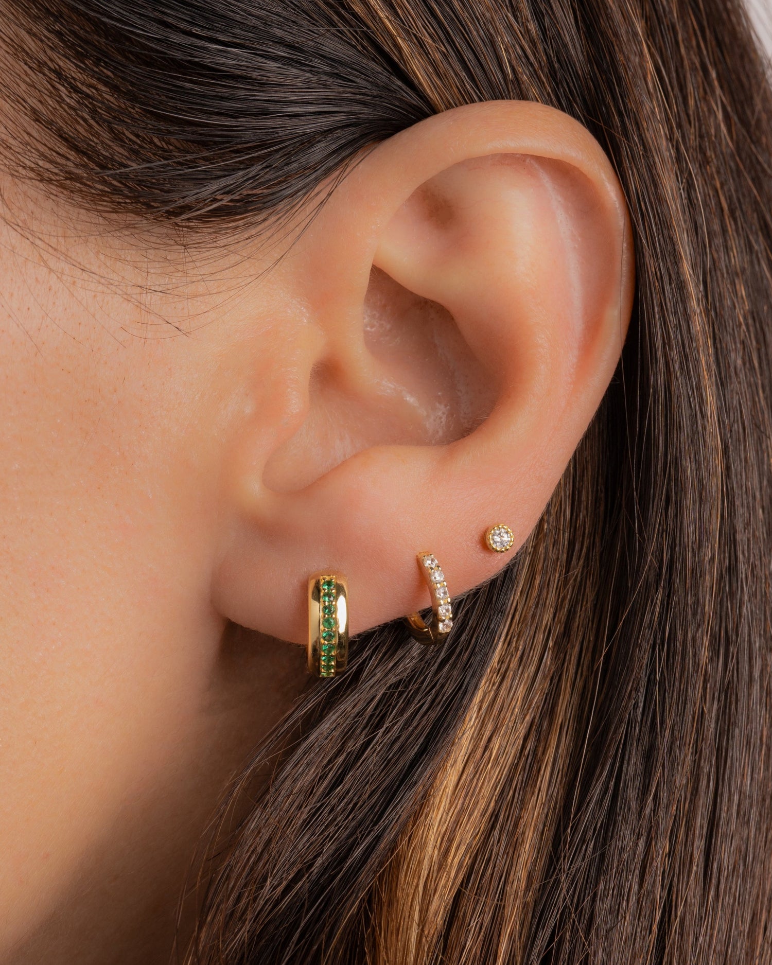 a close up of a model with three piercings, a small gold hoop earring with emerald gemstones in her first piercing, a small diamond huggie hoop in her second piercing, and a tiny diamond stud on her third piercing