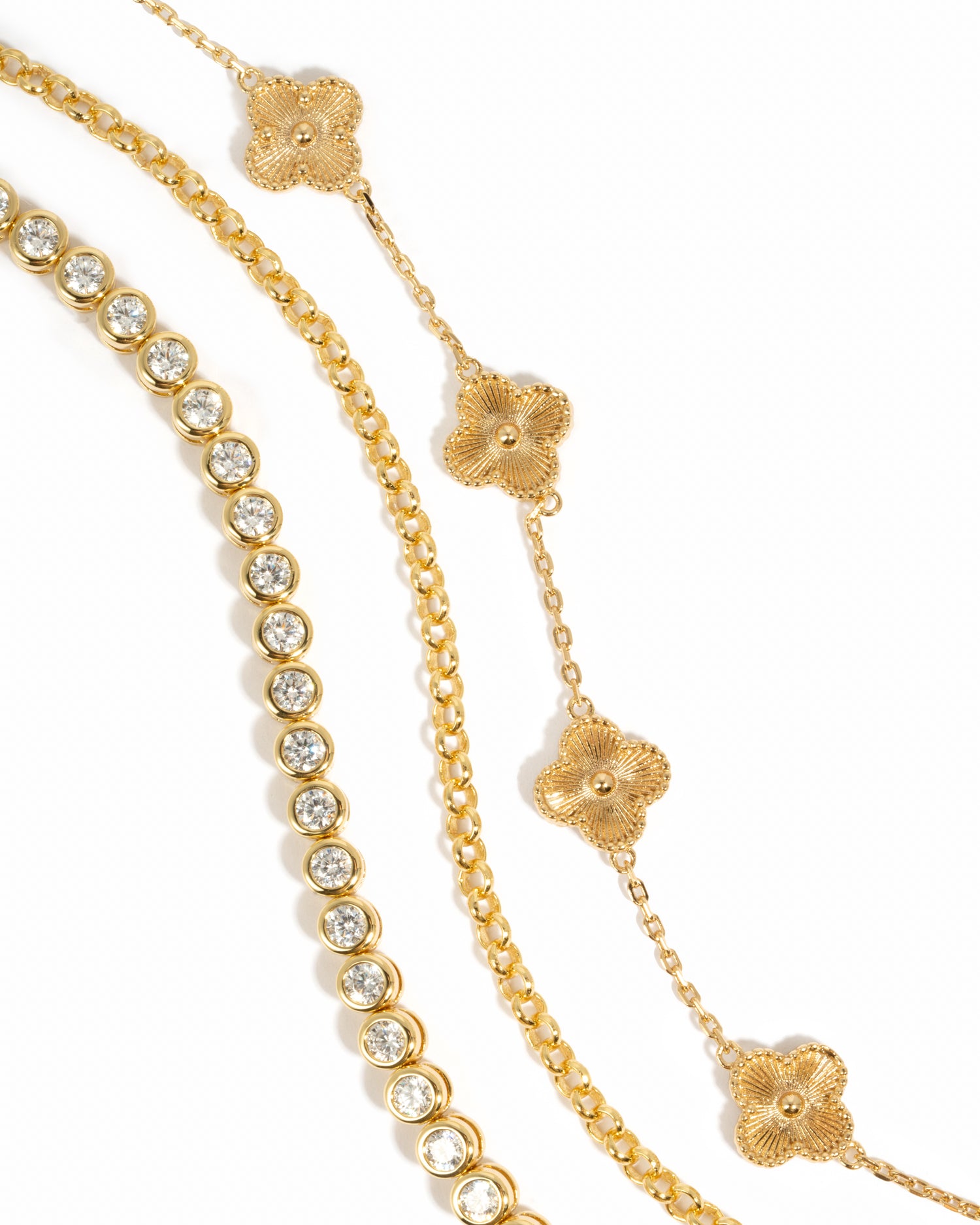 Gold bracelet stack featuring a round bezel tennis bracelet, a simple gold chain bracelet, and a gold chain four leaf clover stackable charm bracelet on a white background