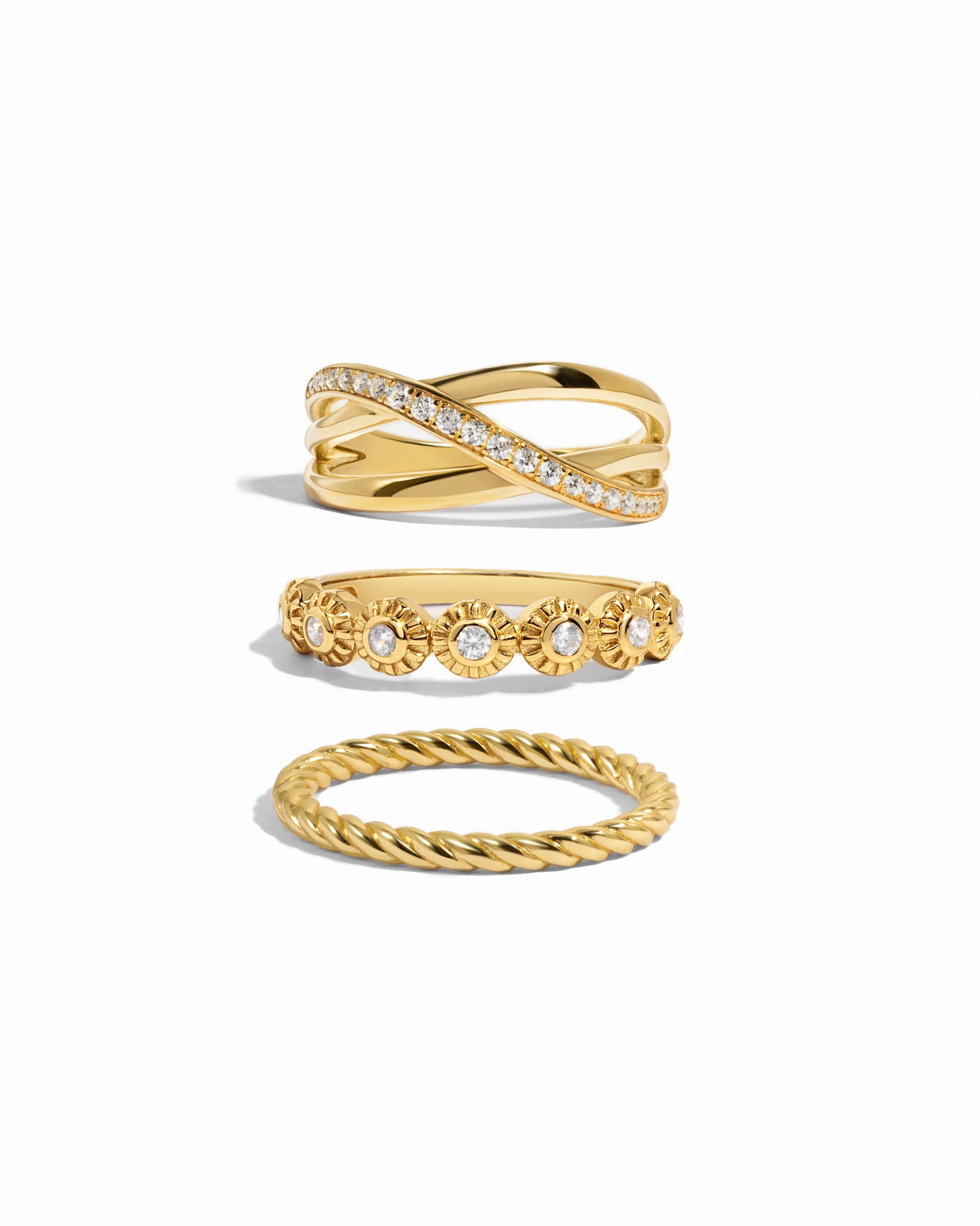 Gold stackable rings including a diamond x band ring with a diamond slash, a gold bezel set textured gold ring, and a thick spiral band ring on a white background