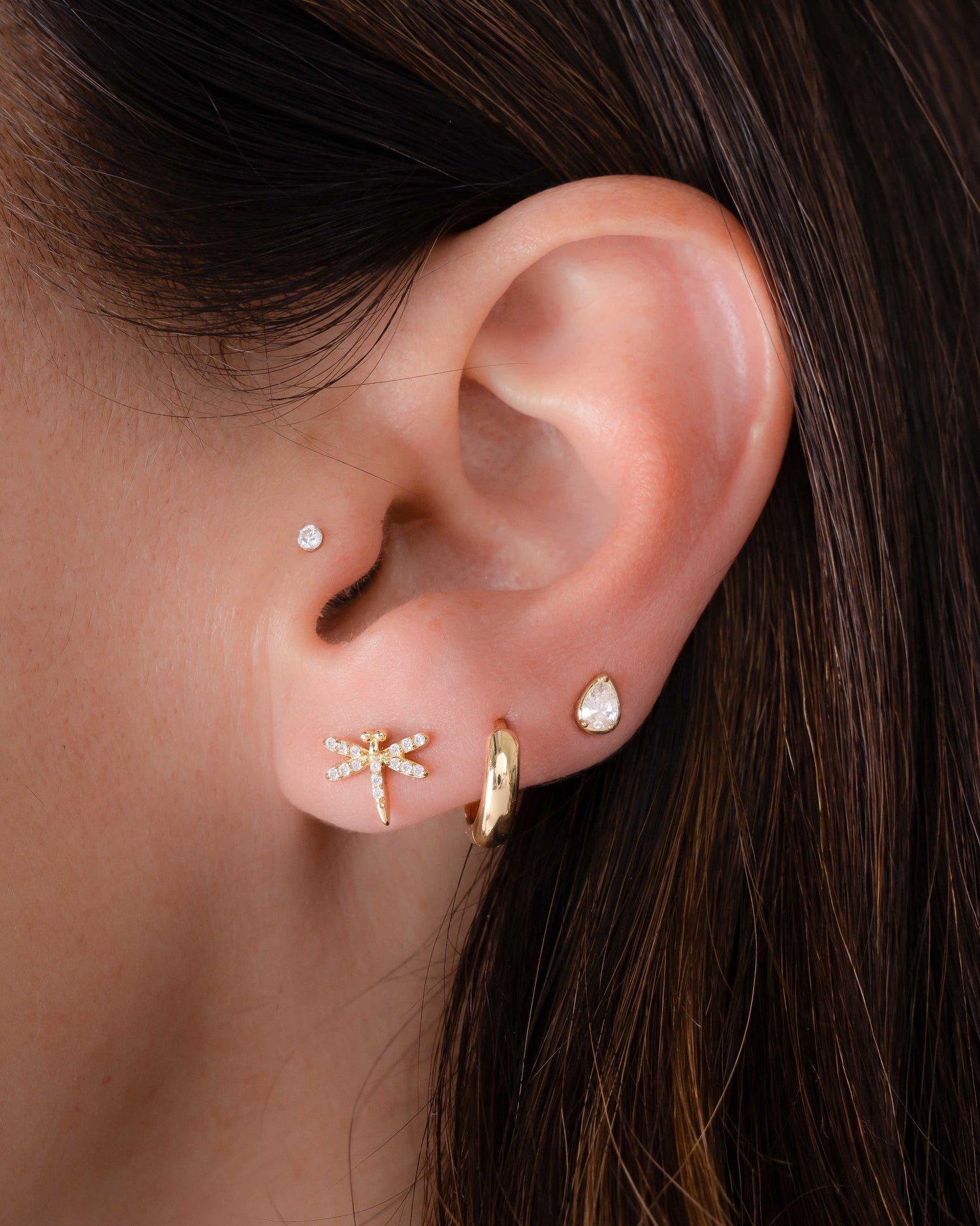 Model wearing gold dragonfly studs with cubic zirconia stones, a minimalist huggie, and a pear stud earring