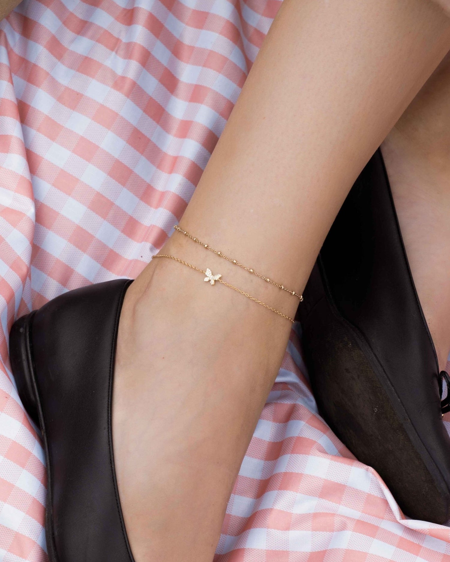 Person sitting on a pink and white checkered blanket with a glass of red wine, holding a bouquet of flowers, wearing black shoes and gold beaded ankle bracelet with a butterfly charm ankle bracelet