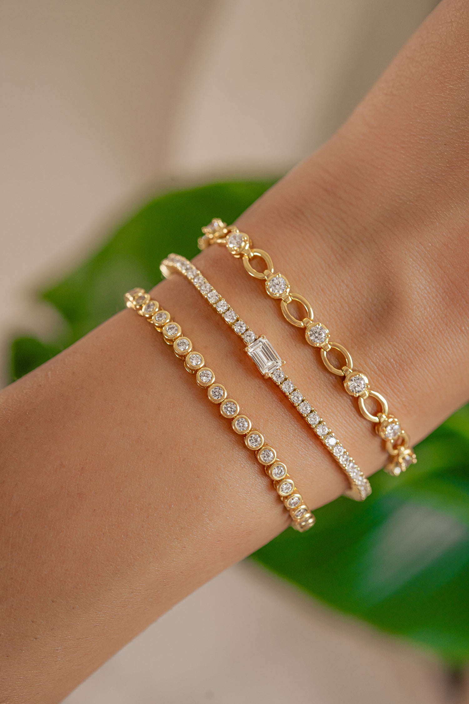 gold bracelet stack featuring a round bezel-set stone bracelet, a cubic zirconia prong bracelet with baguette charm, and a round cz chain tennis bracelet