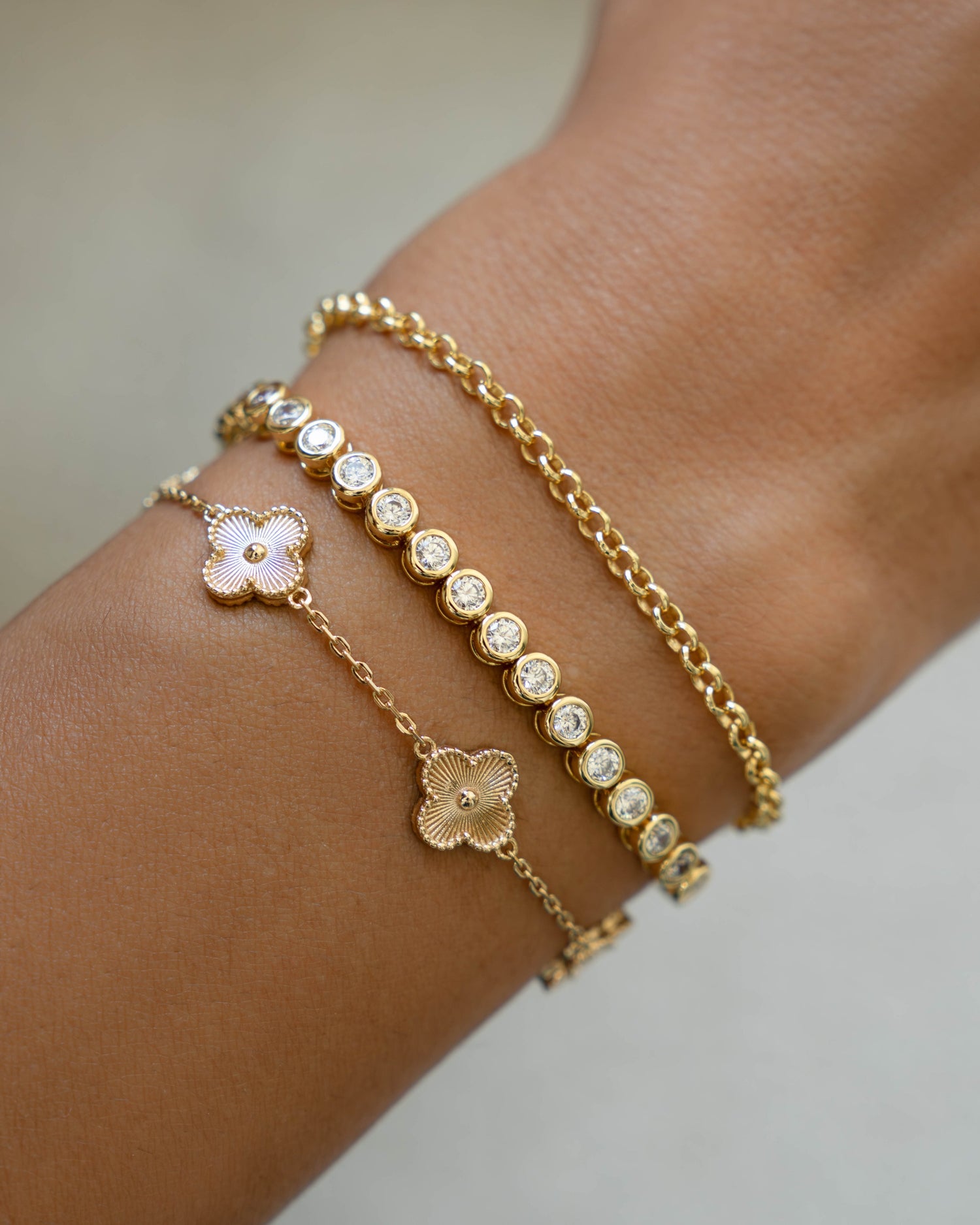 Gold bracelet  stack with four leaf clover design, a simple round bezel tennis bracelet, and a dainty gold chain bracelet on a wrist against a neutral background