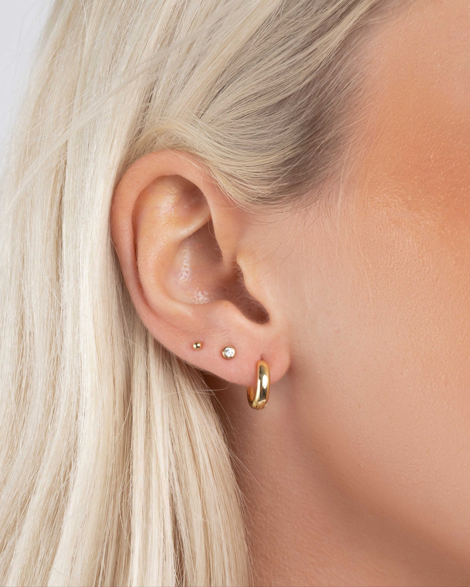 Close-up of an ear with small gold chunky hoop earrings, a diamond stud earring, and a gold ball stud earring on model