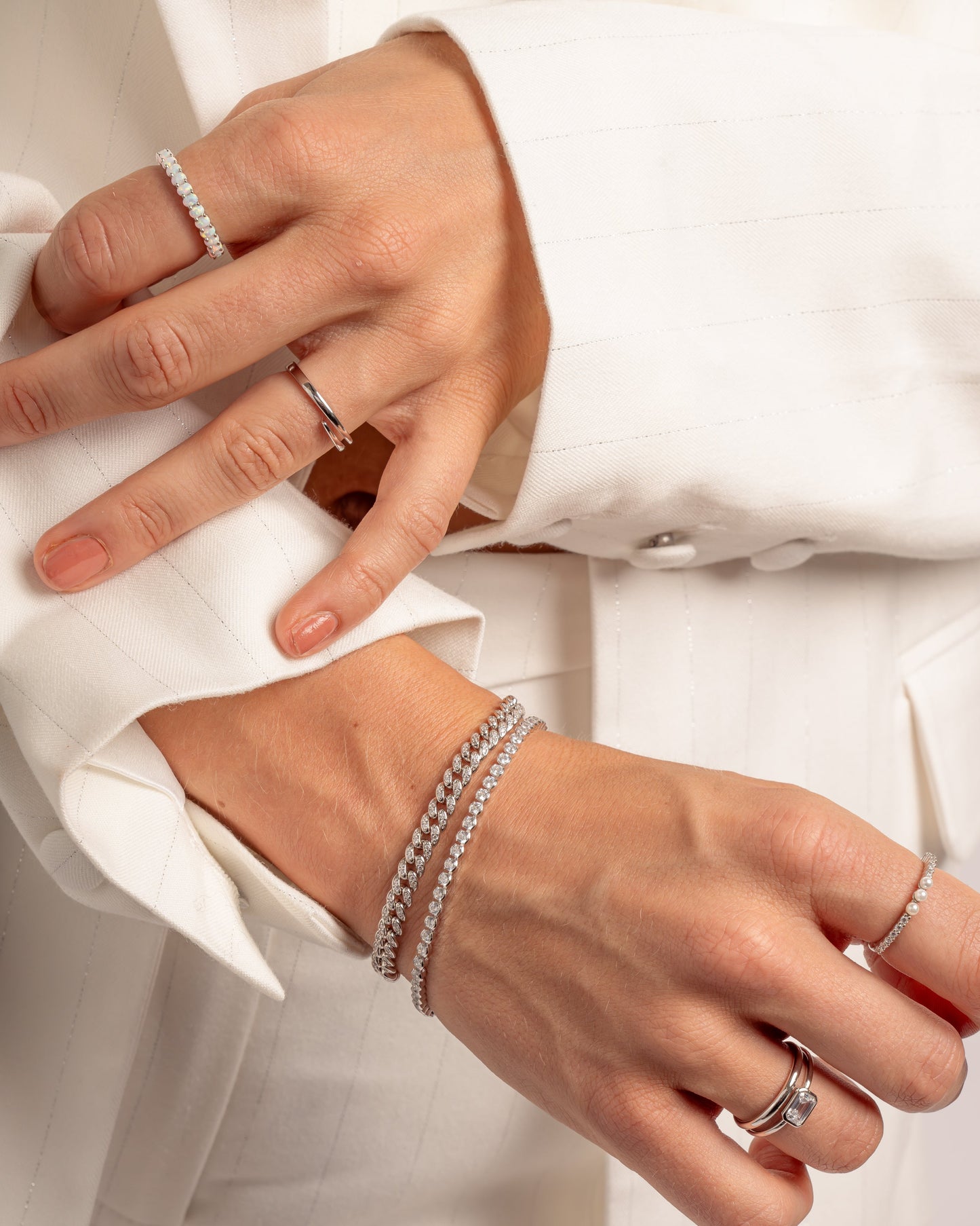 model wearing silver jewelry featuring open spiral ring, opal eternity ring, three pearl ring, and simple baguette ring. Model is also wearing a silver pave cuban chain bracelet, and a 3mm round tennis bracelet
