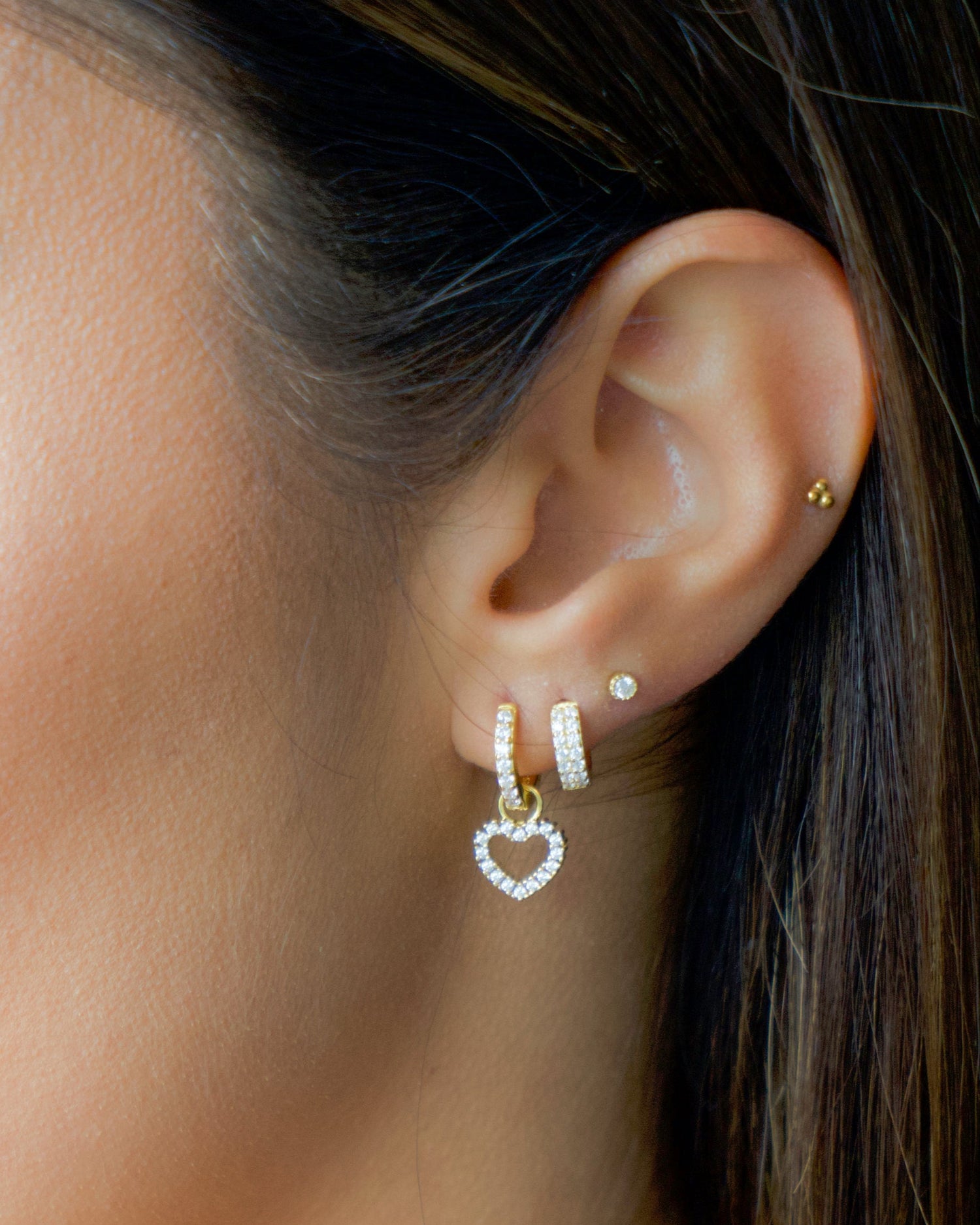 Model wearing pavé feminine gold earring stack including a diamond stud, a diamond huggie, and a heart charm diamond hoop.