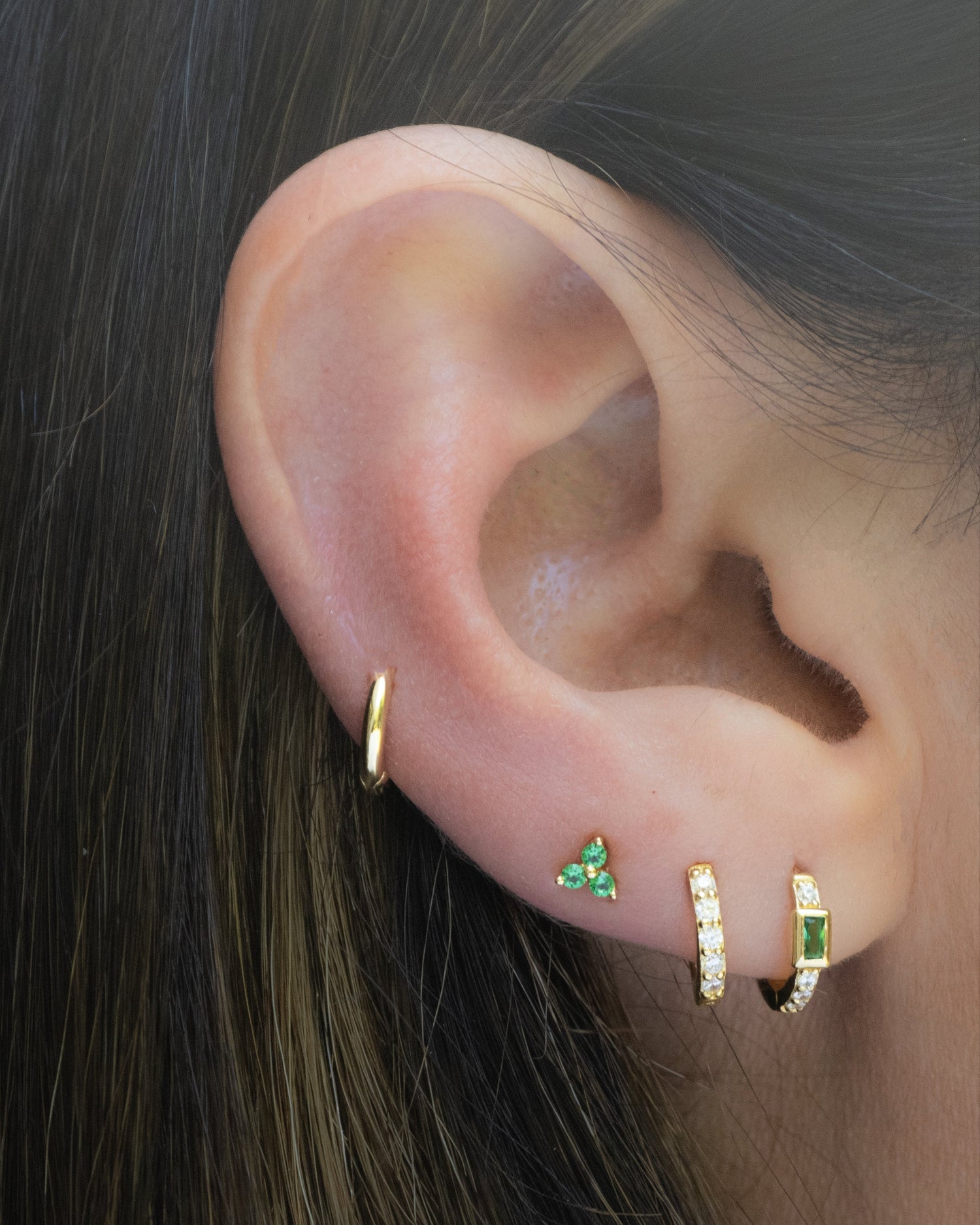 emerald earring stack featuring a plain huggie, a emerald 3 stone stud earring, a cz huggie earring, and a emerald baguette huggie earring
