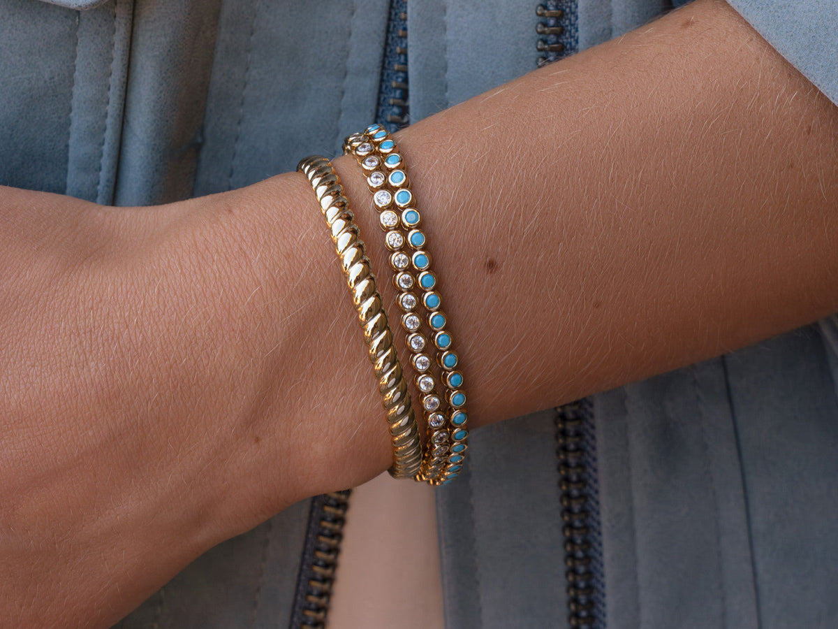 Three gold-tone bracelets with clear stones, turquoise color and a rope cuff, worn with a blue jacket.