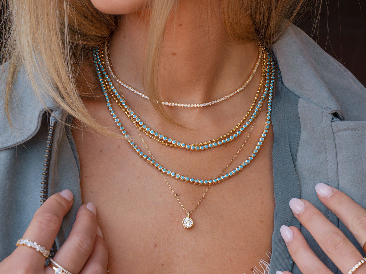 Turquoise necklace layers, a clear tennis necklace, a textured gold-tone chain and a round bezel pendant, worn with a blue jacket.