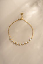 A delicate gold chain bracelet featuring a row of small, pear-cut gemstones, displayed on a soft beige background with a warm, sunlit shadow overlay.