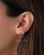 Close-up of an ear wearing a 7mm diamond huggie hoop earring layered with an 8mm diamond huggie with text indicating gold content and size.