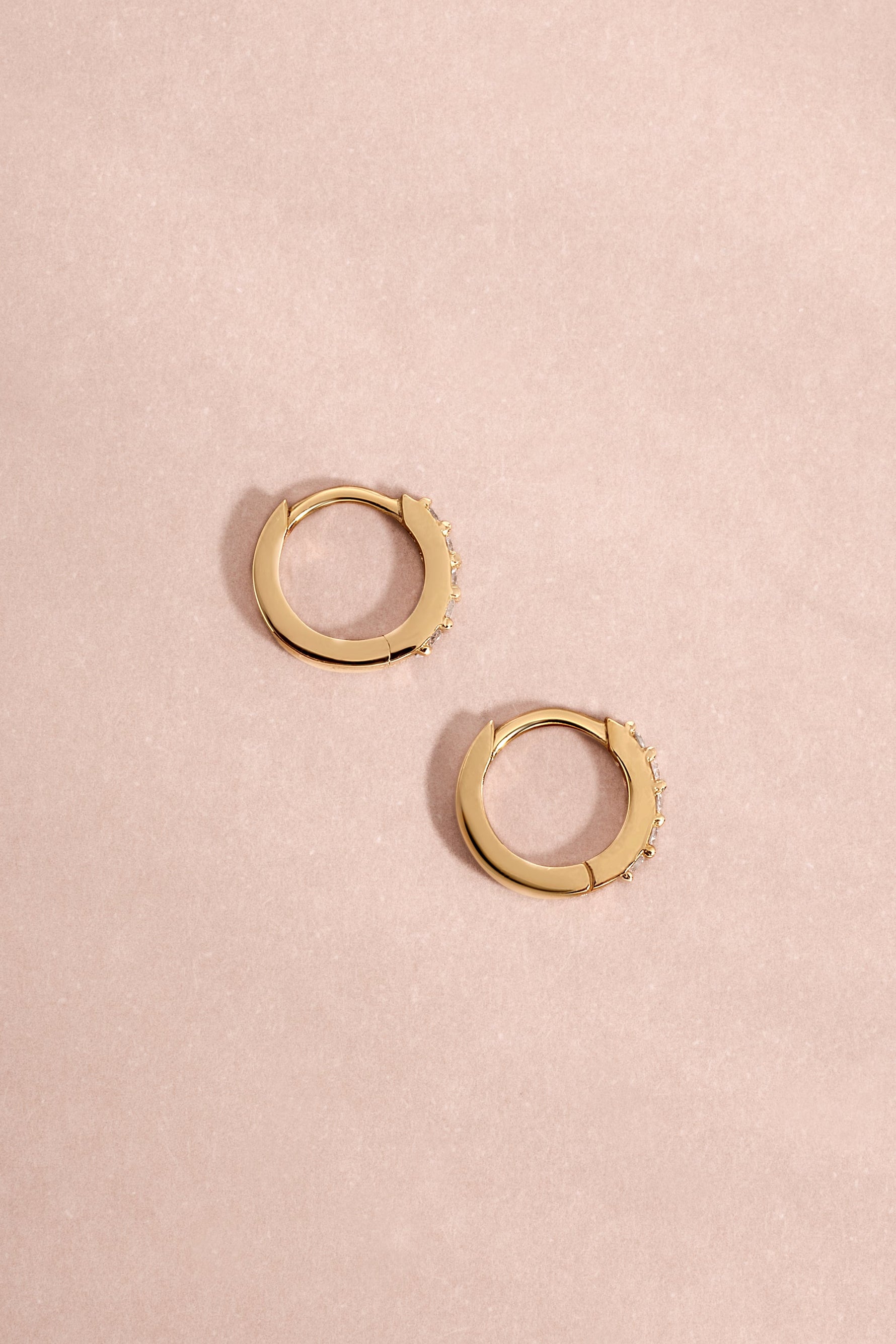 Pair of 14K solid gold 7mm diamond huggie earrings flat lay on soft pink background