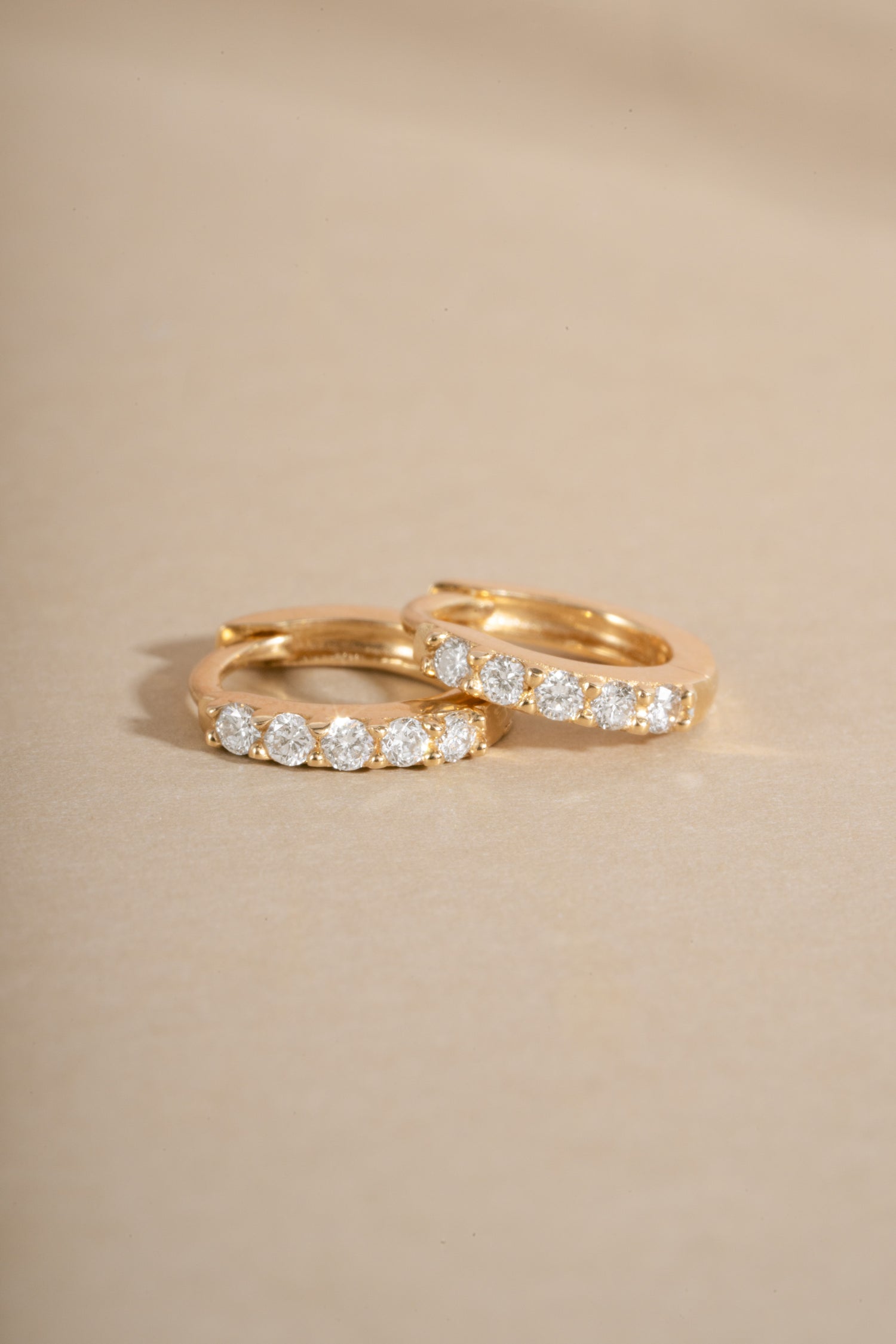 Two gold huggie hoop rings, each set with a row of round clear gemstones, resting on a warm beige background.