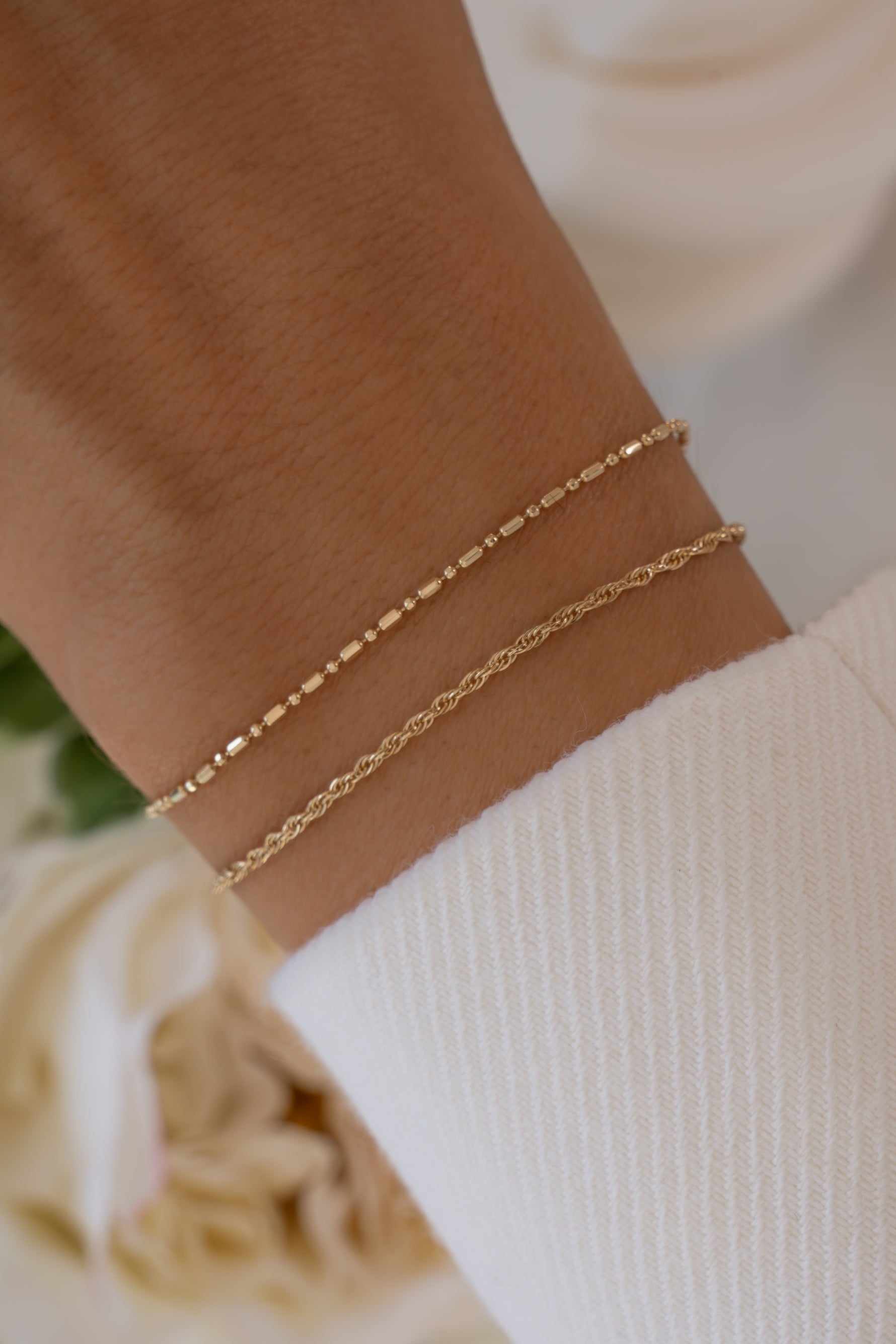 14K solid gold bar station chain bracelet layered with rope chain bracelet worn on wrist with blurred florals in background