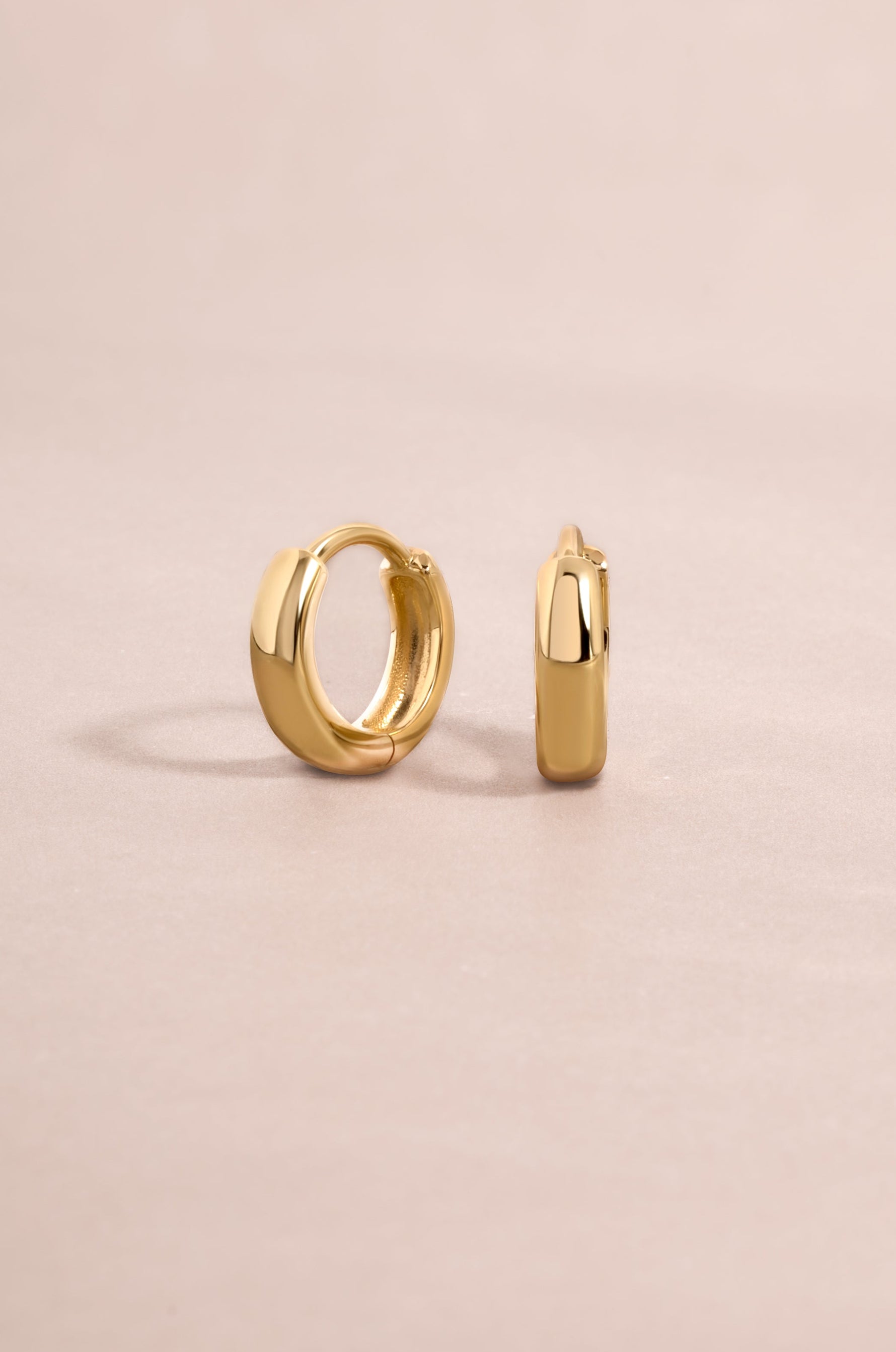 14K solid gold chunky huggie earrings by Sami Jewels displayed on a pink background.