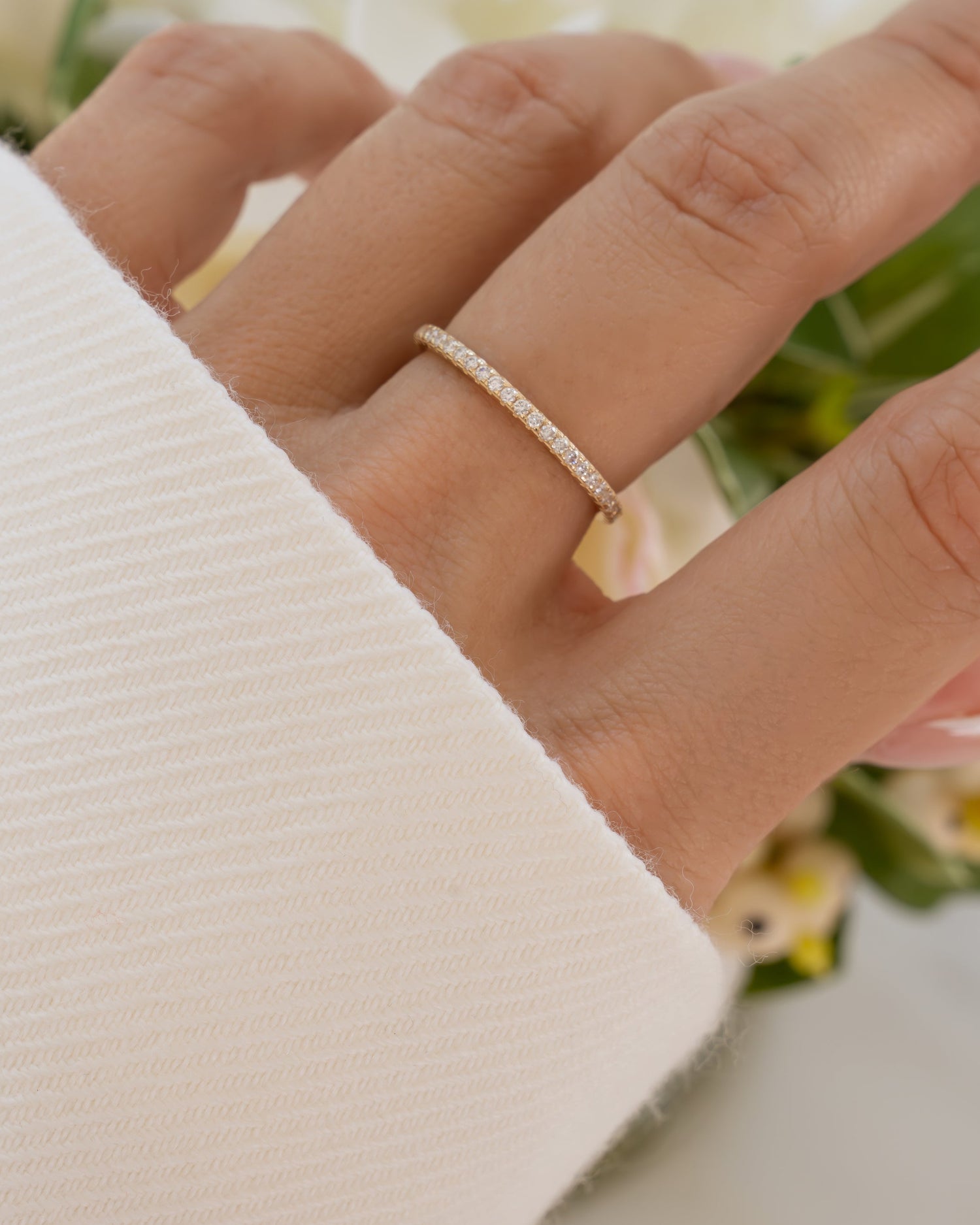 Close-up of a hand wearing a 14K solid gold pave eternity ring with a blurred floral background