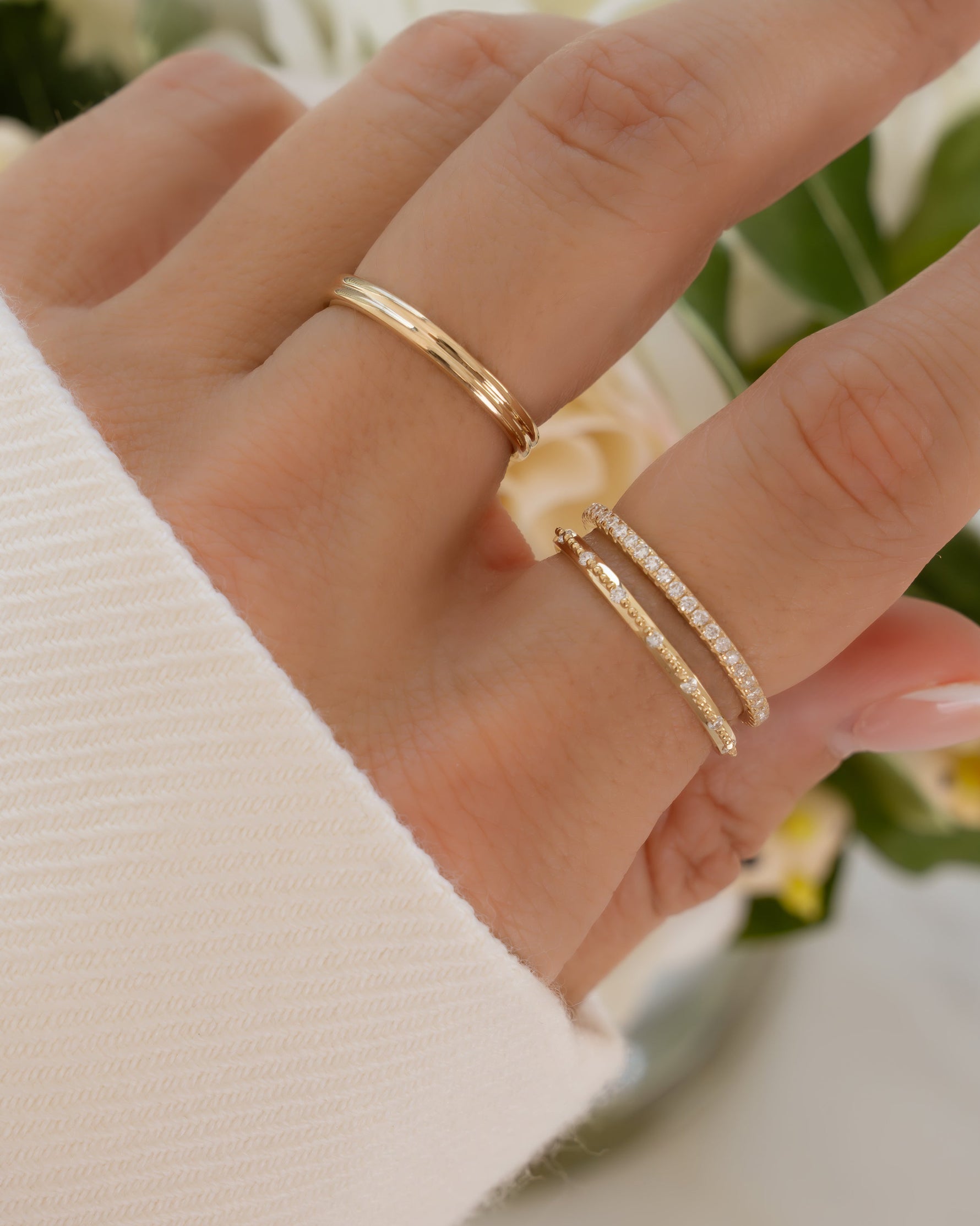 Hand wearing stackable diamond gold rings crafted in 14k solid gold and lab grown diamonds with a blurred floral background