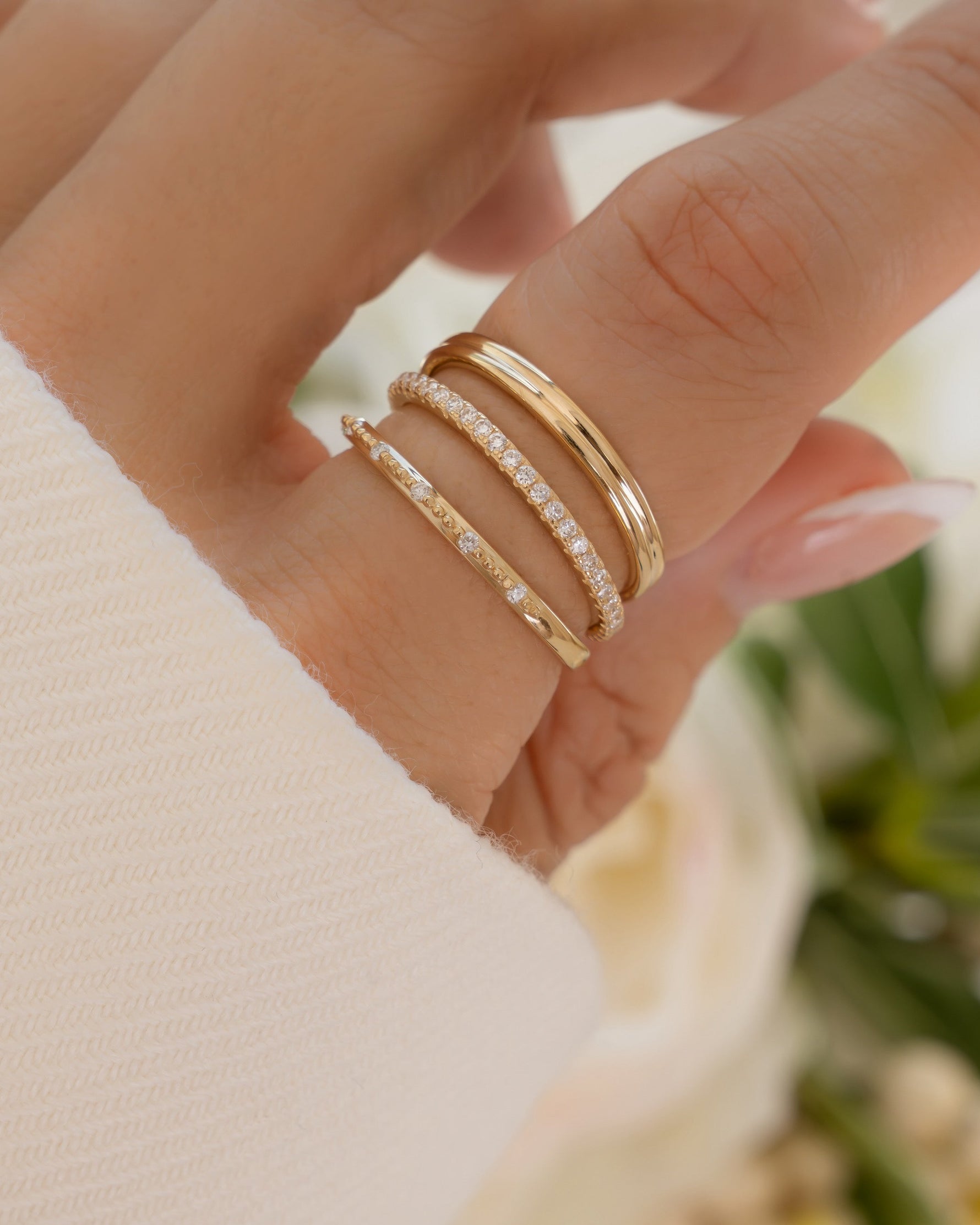 Close-up of hand wearing dainty 14K solid gold diamond stacking rings with lab grown diamonds with blurred floral background
