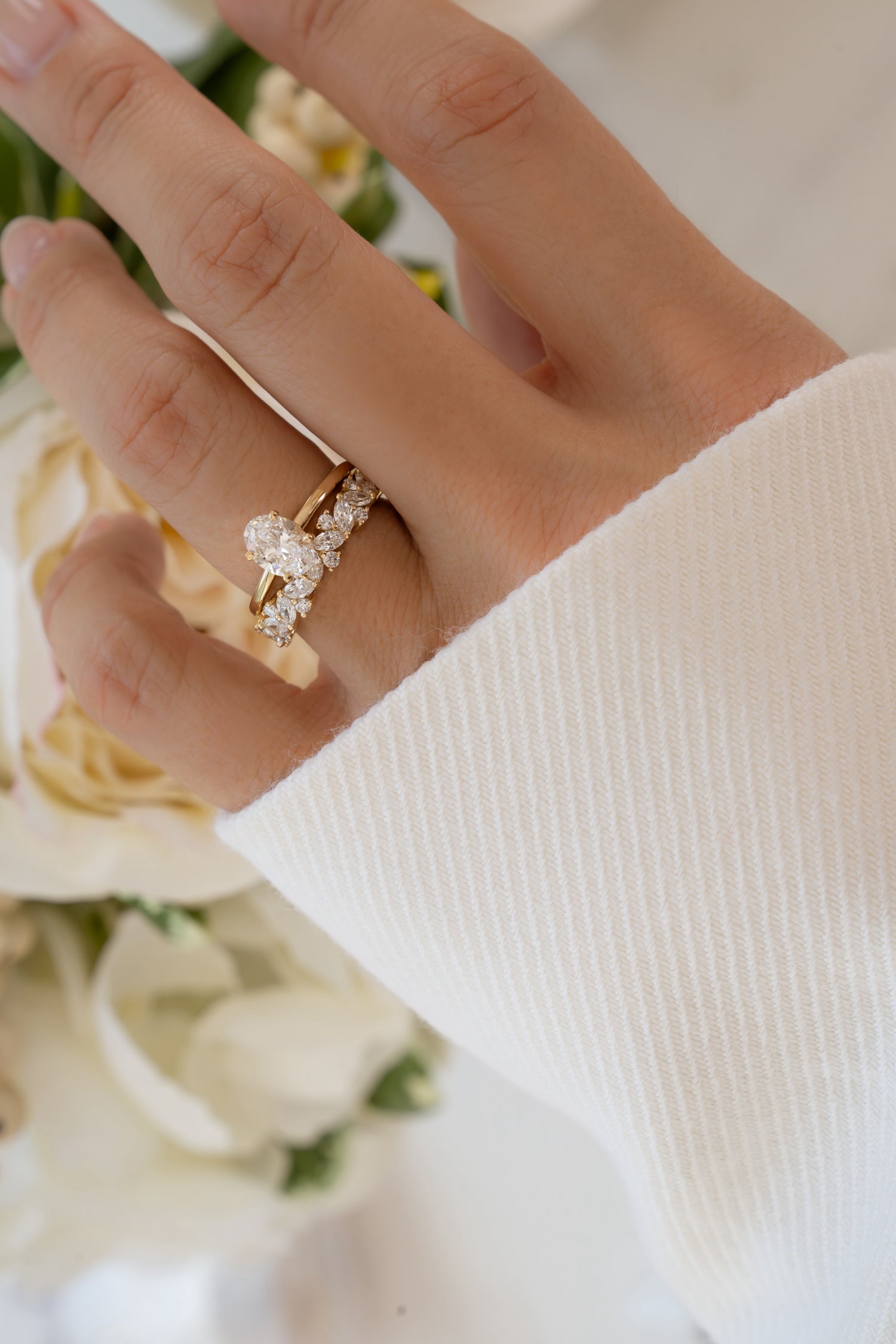 Close-up of a hand wearing a solid gold ring with a oval diamond paired with a multi stone diamond engagement ring, against a blurred background of white flowers.