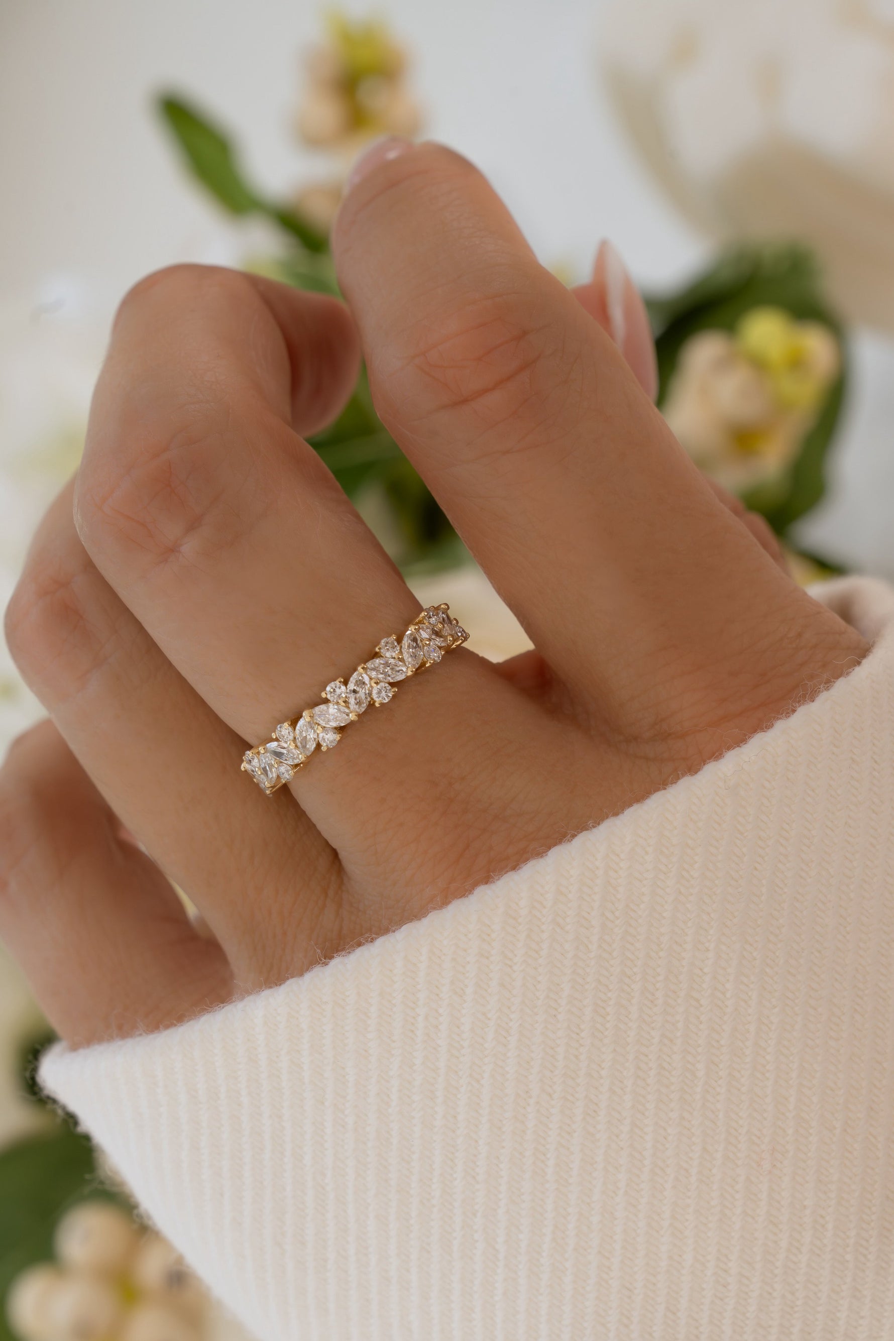 A close-up of a hand wearing a delicate diamond eternity ring on a gold band, styled with a white ribbed knit sleeve against a soft, blurred background of white flowers.