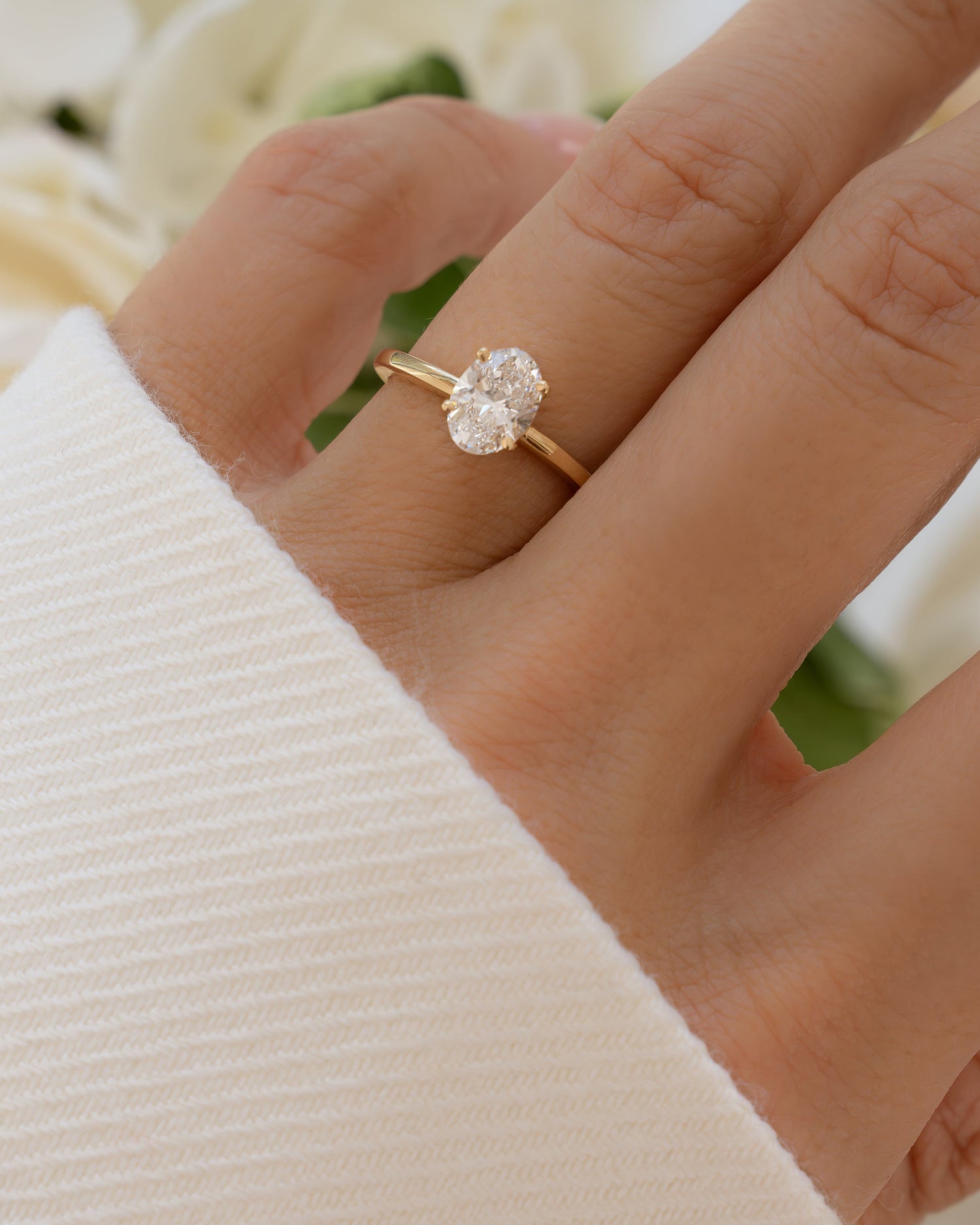 Solid 14k yellow gold oval solitaire ring featuring a lab grown diamond worn on hand with blurred floral background