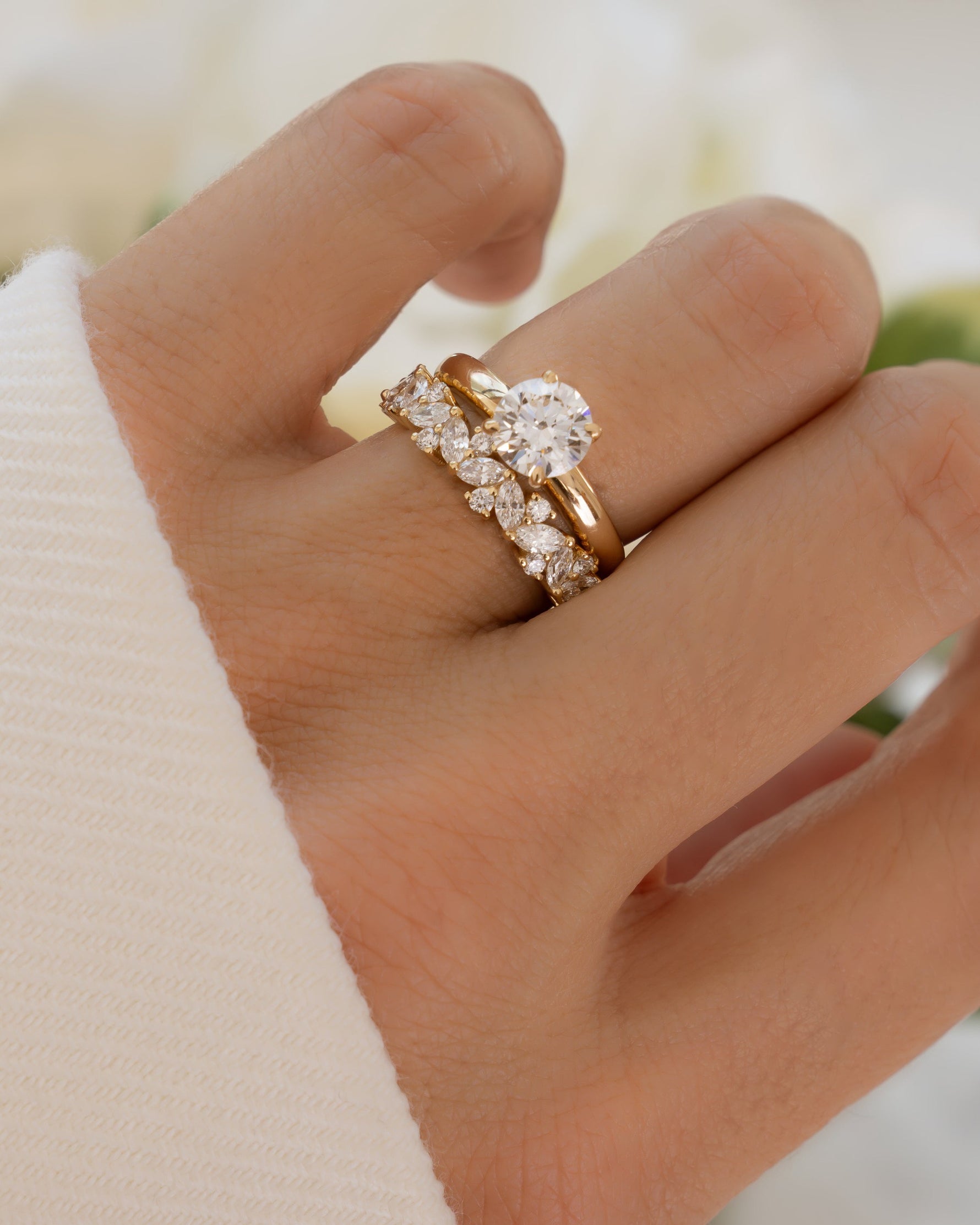 Hand wearing a 14K solid gold ring with a multi stone diamond engagement band against a blurred floral background