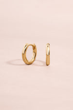 Minimalist solid gold huggie earrings on a light pink background