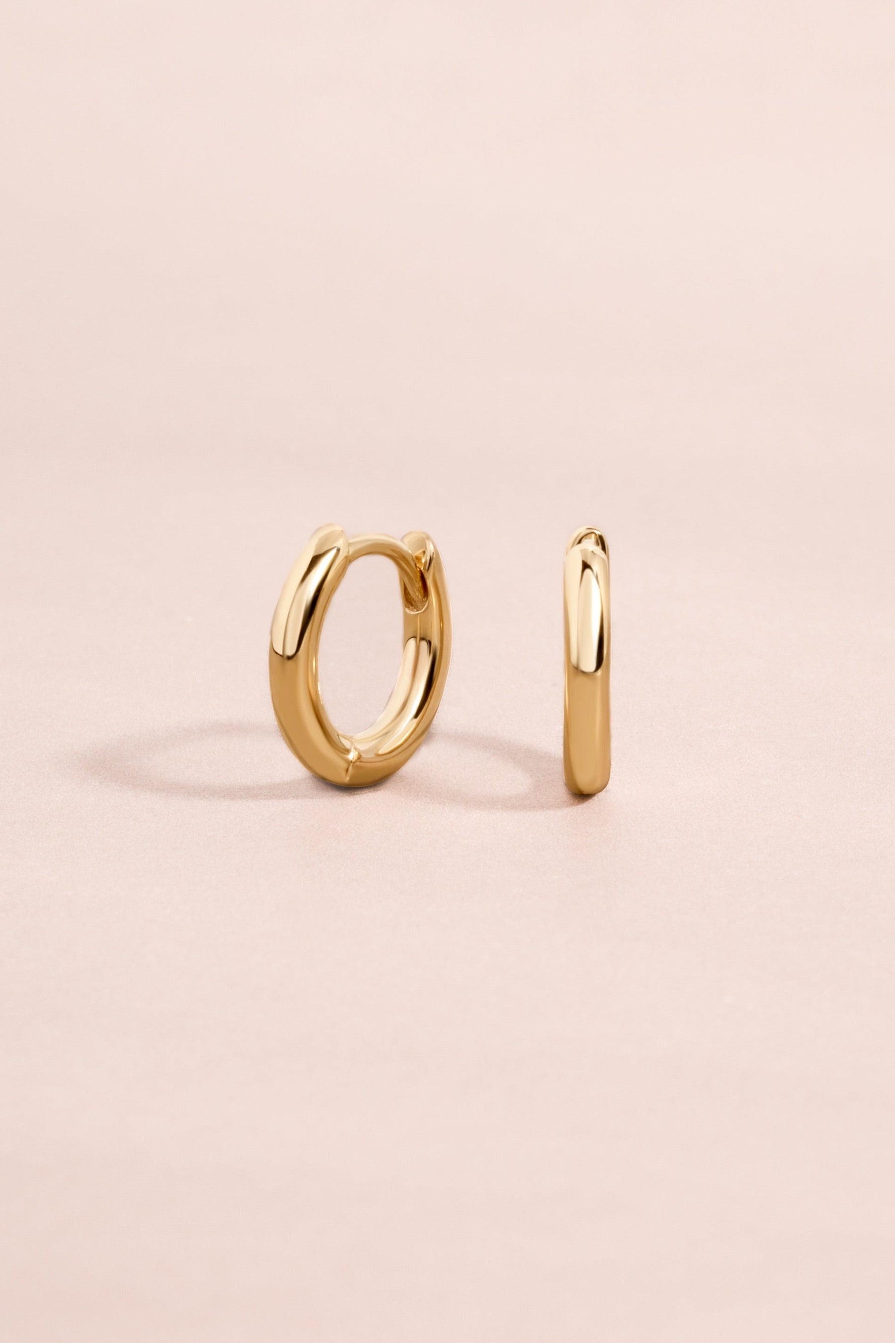 Minimalist solid gold huggie earrings on a light pink background