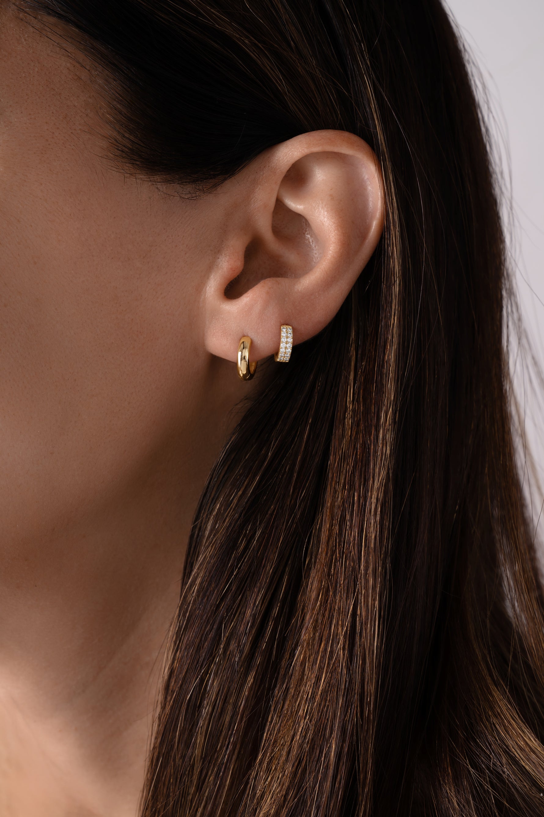 Close-up of a person wearing gold hoop earrings stacked with diamond huggies with a neutral background