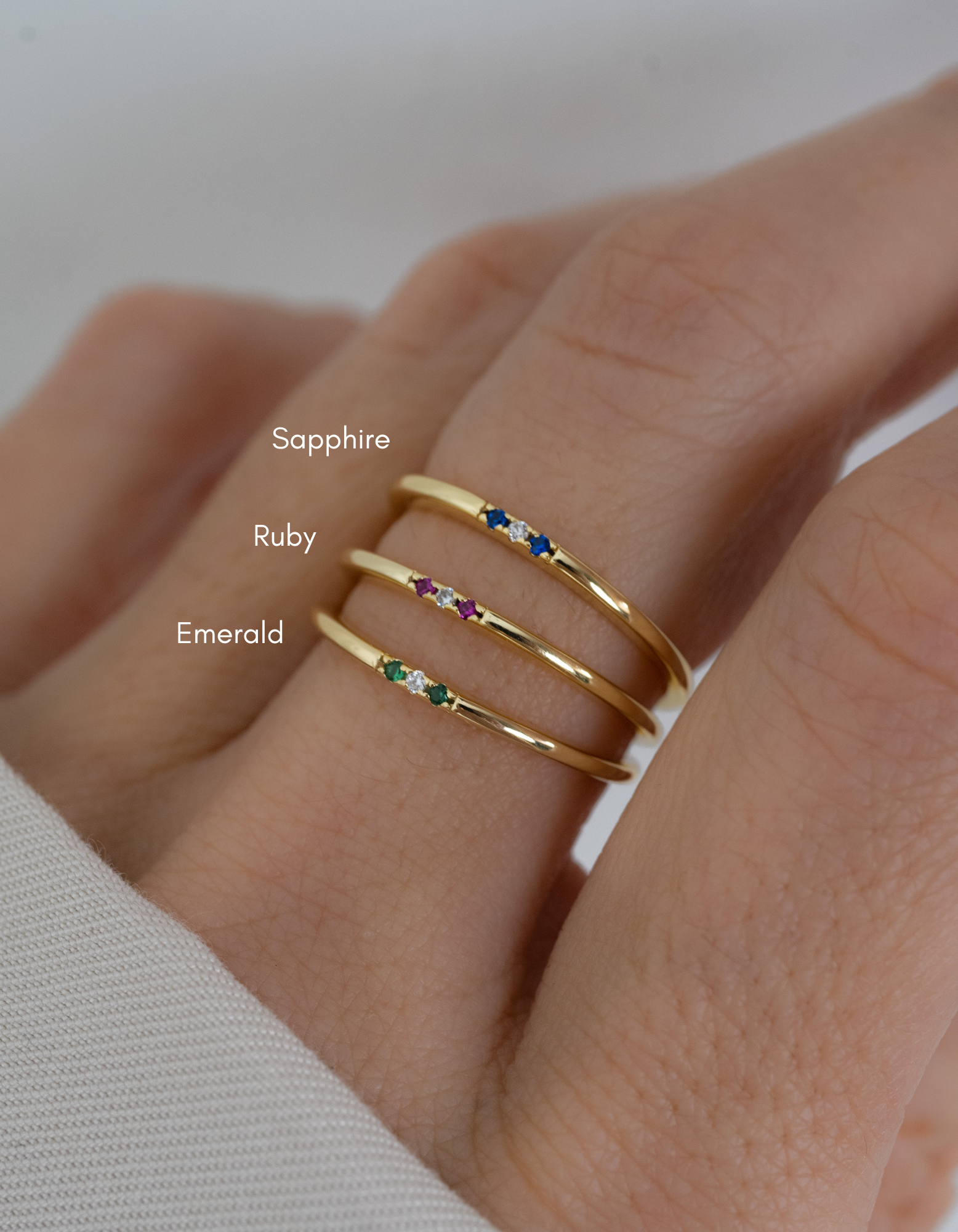 Three gold 3 stone gemstone rings in sapphire, ruby and emerald on a finger against a neutral background