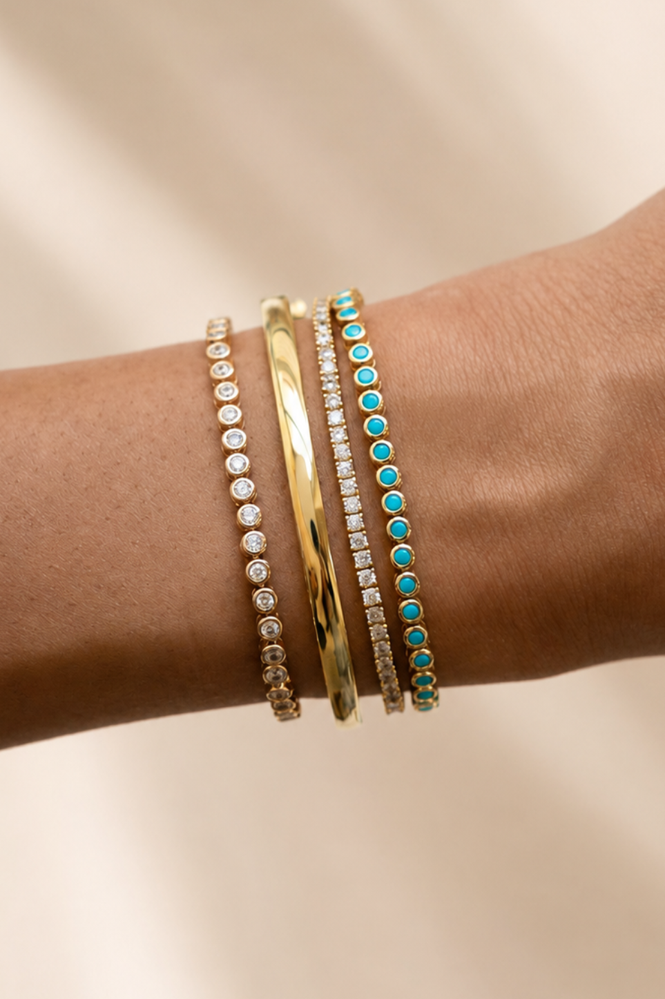 A close-up of a person's wrist wearing three slim bracelets: two gold-toned bracelets, one with clear stones and one with turquoise stones.