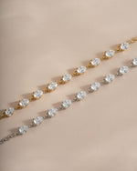 Two tennis-style bracelets with pear-cut clear stones, one in yellow gold and one in silver, displayed diagonally on a warm beige background.