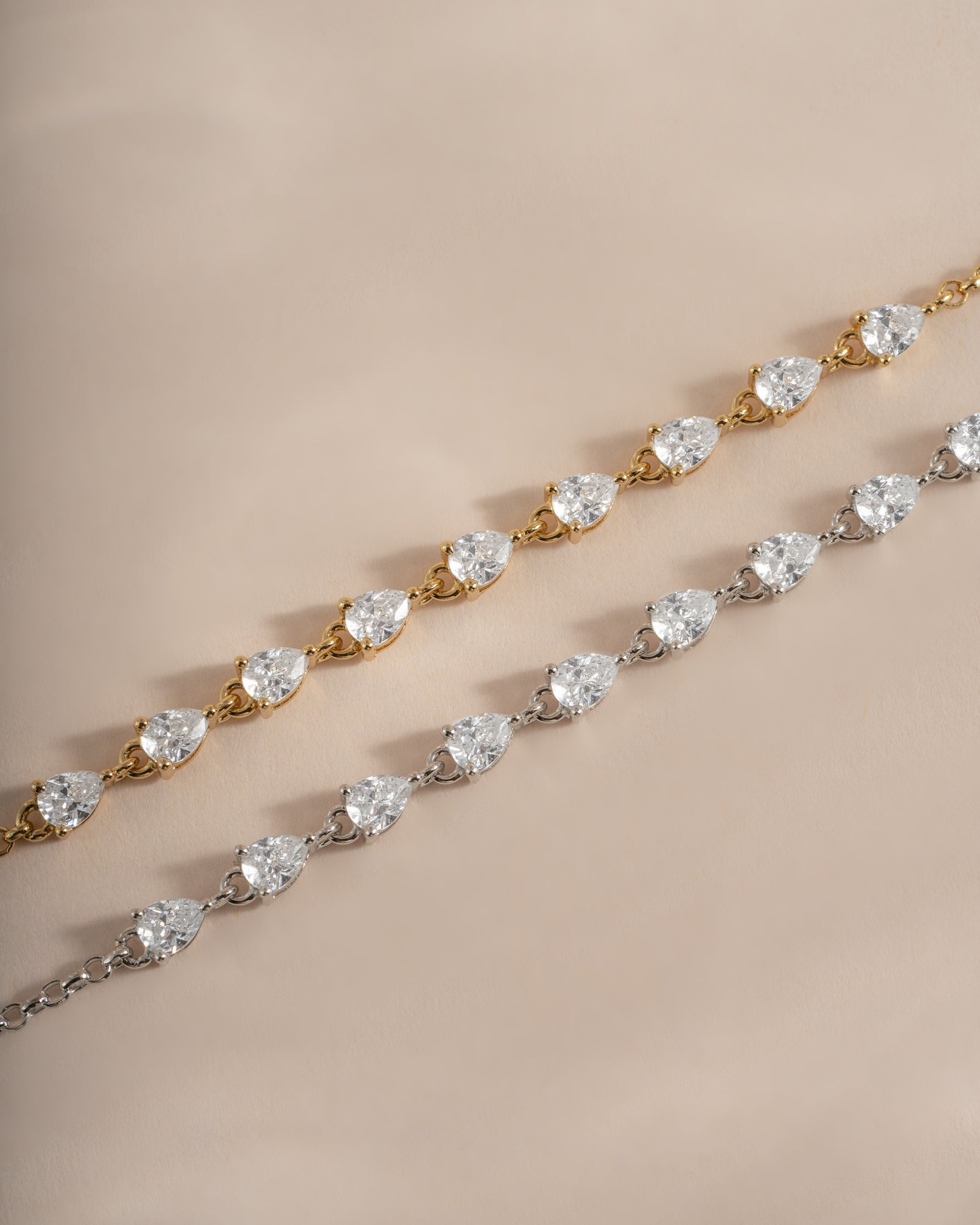Two tennis-style bracelets with pear-cut clear stones, one in yellow gold and one in silver, displayed diagonally on a warm beige background.