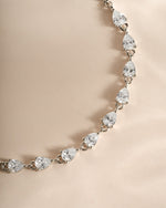 A close-up of a silver-toned tennis bracelet featuring pear-cut clear gemstones, displayed on a soft beige satin background.