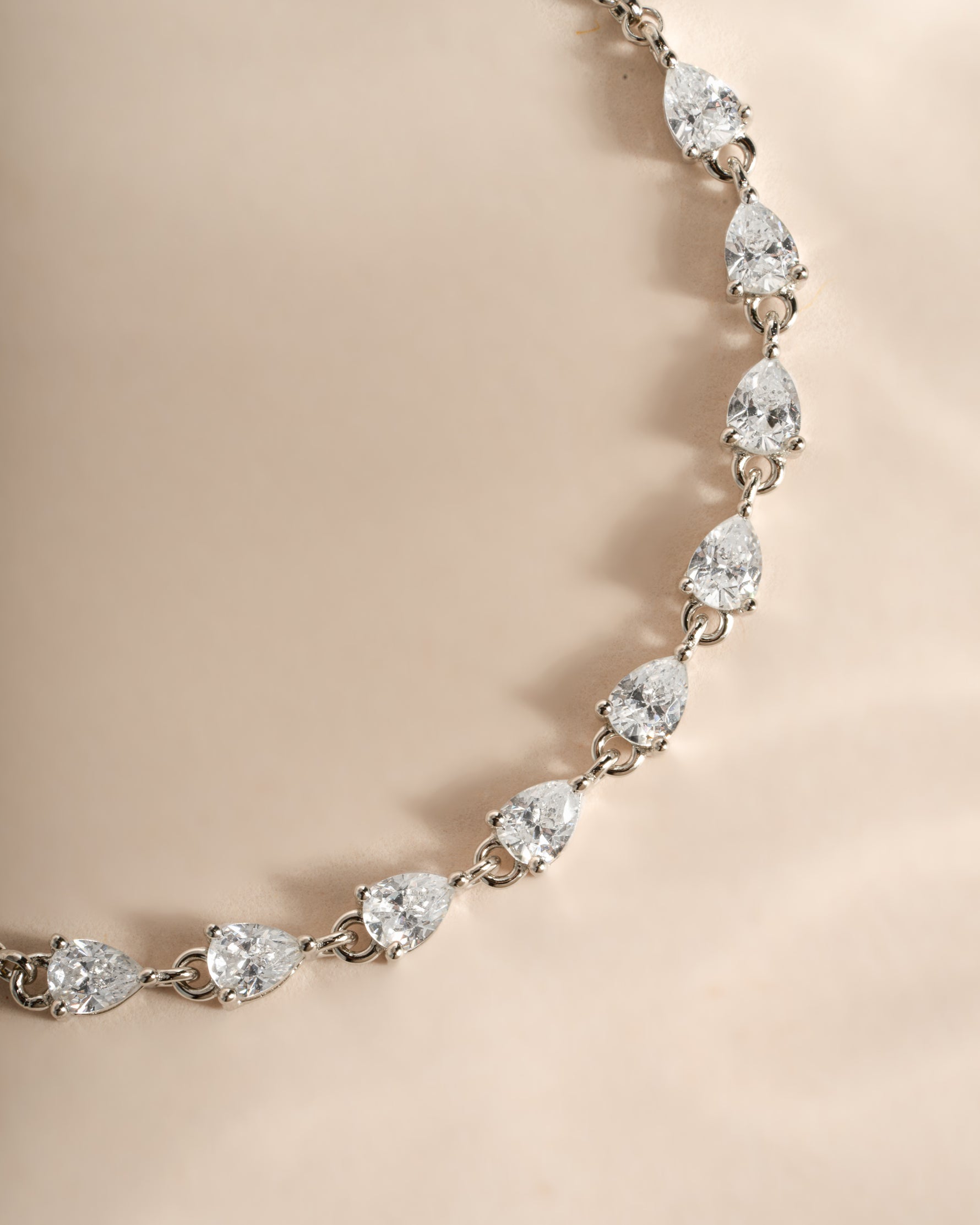 A close-up of a silver-toned tennis bracelet featuring pear-cut clear gemstones, displayed on a soft beige satin background.