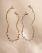 Two delicate chain bracelets with diamond teardrop crystals, one in gold-tone metal and one in silver-tone metal, displayed on a soft beige background.