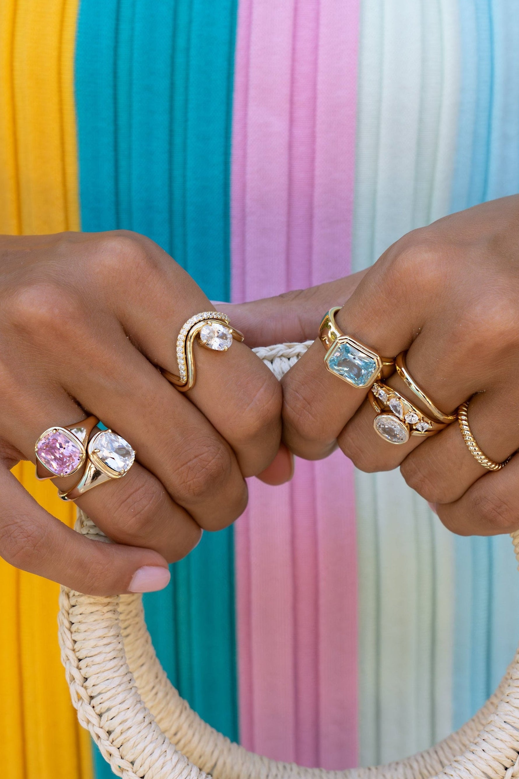 Close-up of two hands wearing multiple gold rings with colorful gemstones, holding the handle of a woven rattan handbag against a pastel striped background.