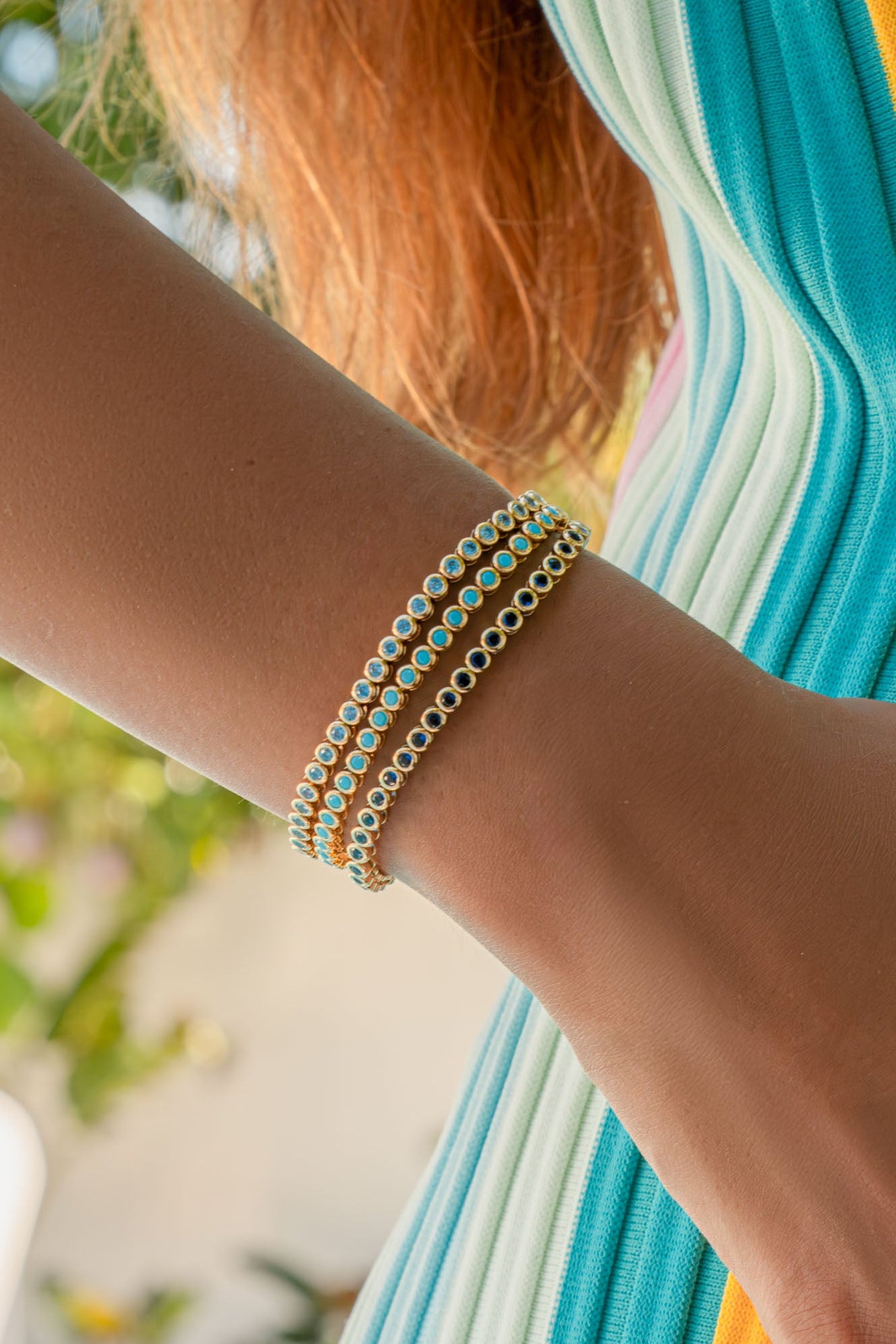 A close-up of a person's hand wearing gold rings and a triple-strand beaded bracelet, resting on a vibrant, vertically-striped garment with yellow, pink, blue, and white colors.