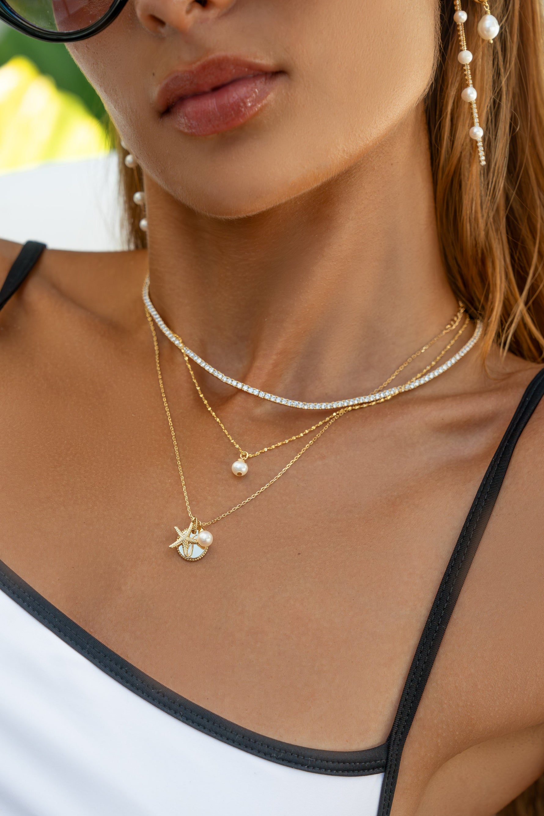 Close-up of a woman wearing layered gold necklaces, including a pearl drop necklace and a small shell pendant, along with dangling pearl earrings and a white top with black trim.