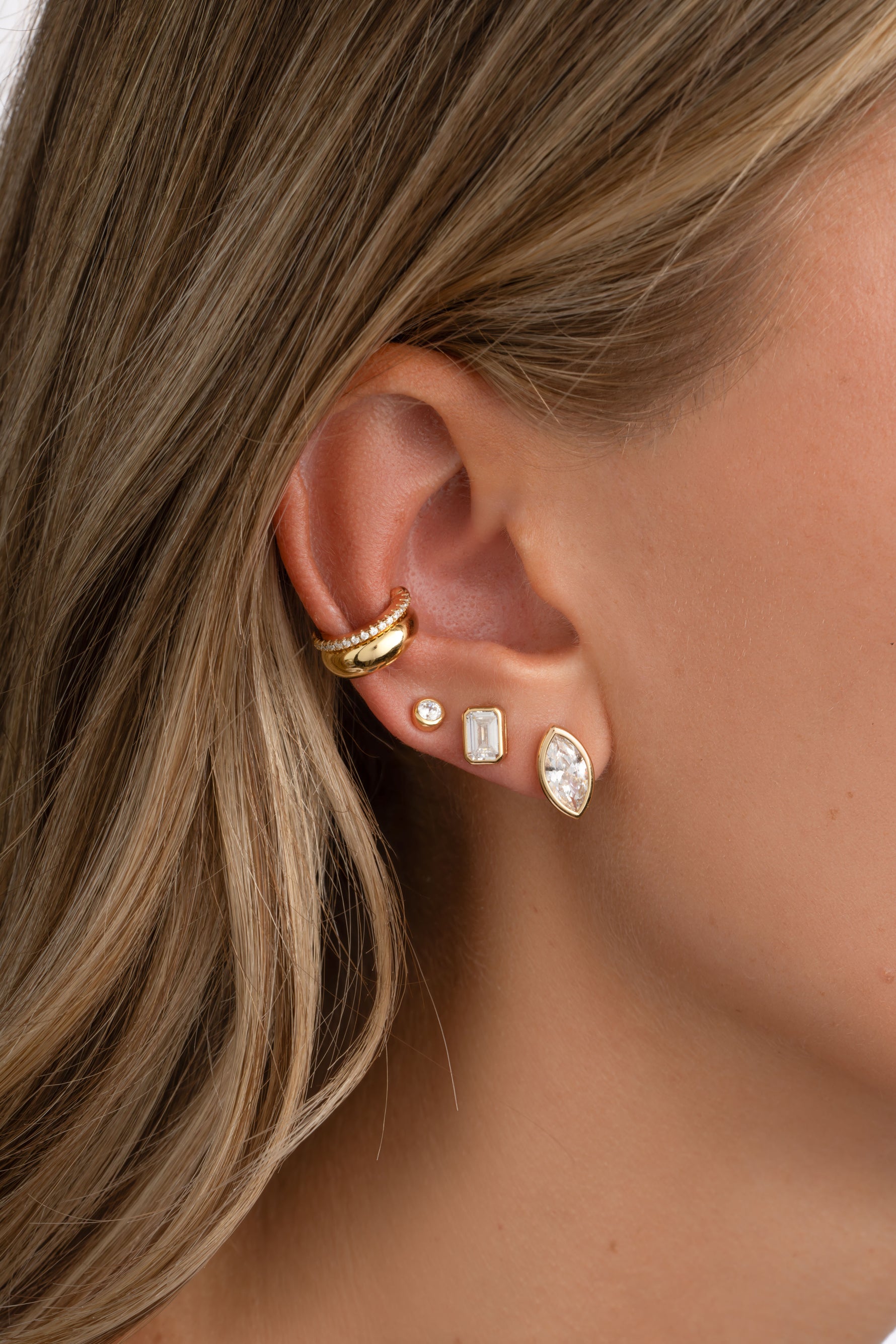 A close-up of a model's ear showcasing a stack of gold ear piercings, including a gold hoop cartilage piercing, a small gold stud, a rectangular gold stud, and a gold teardrop stud with a clear marquise-cut gemstone.
