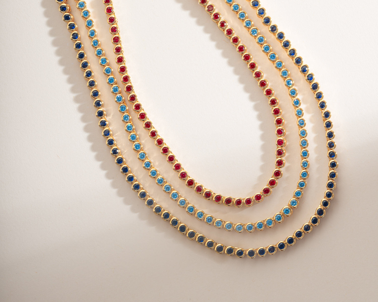 Three gold tennis-style necklaces draped in parallel loops, each set with small round stones in different colors: blue, red, and dark navy, on a clean off-white background.