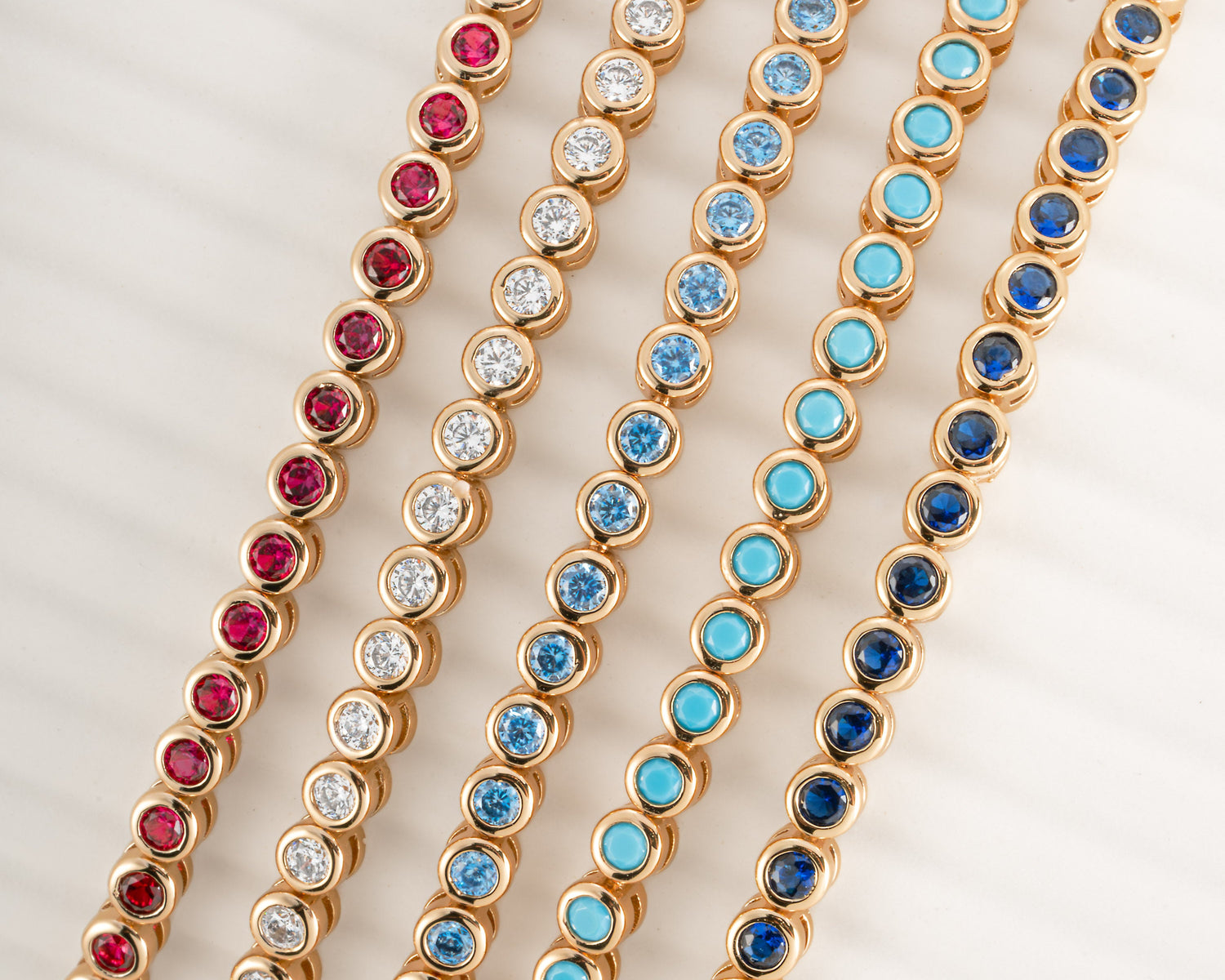 Five gold tennis-style chains arranged parallel to each other on a white background, featuring different colored round stones: red, clear, light blue, medium blue, and dark blue.