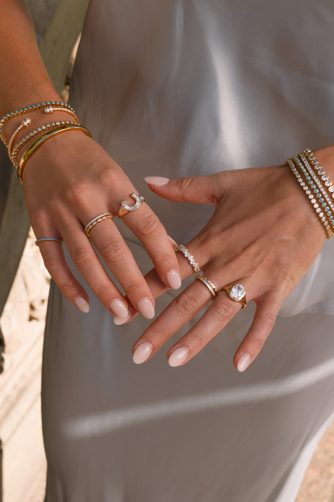 A close-up of a woman's hands wearing multiple delicate rings and stacked bracelets, styled with a light gray satin dress and a dark lace neckline.