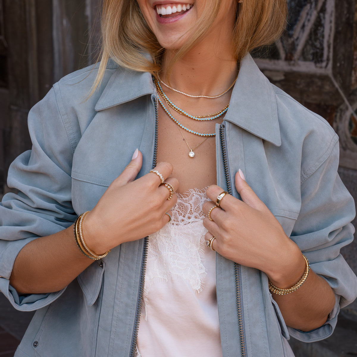 A woman wearing a light blue oversized button-up shirt over a pale pink lace top, accessorized with layered gold necklaces and bracelets, photographed outdoors against a stone wall.