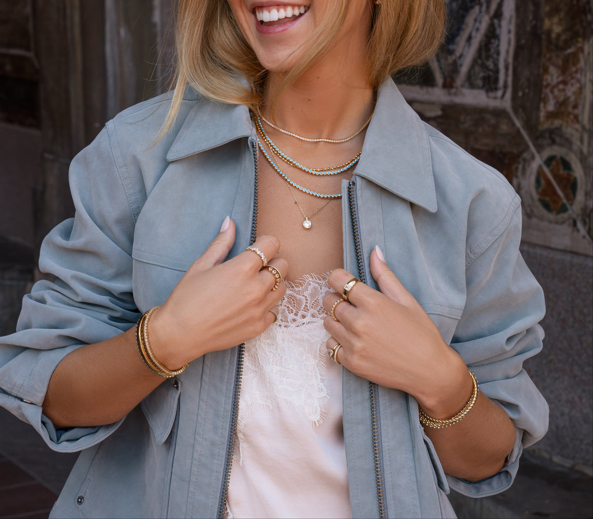 A woman wearing a light blue oversized button-up shirt over a pale pink lace top, accessorized with layered gold necklaces and bracelets, photographed outdoors against a stone wall.
