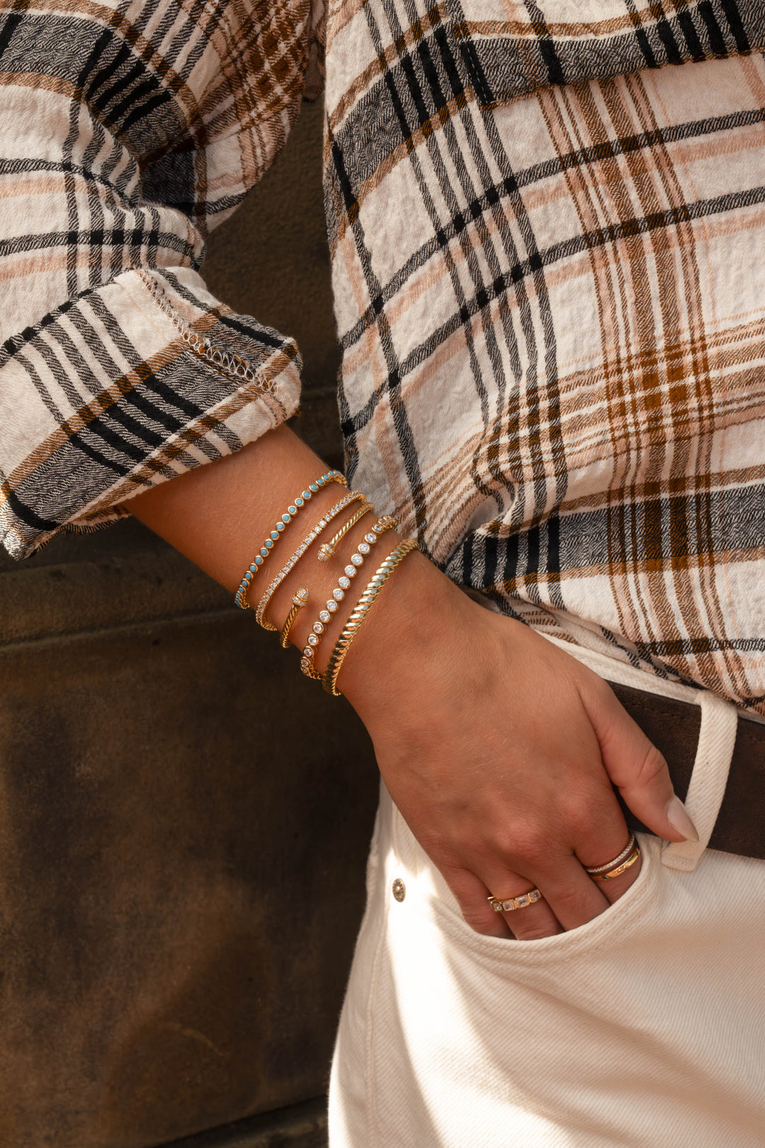 A close-up lifestyle shot of a person wearing a beige, brown, and black plaid linen shirt, cream-colored pants, a dark brown leather belt, and multiple gold bracelets and rings on their wrist and hand.