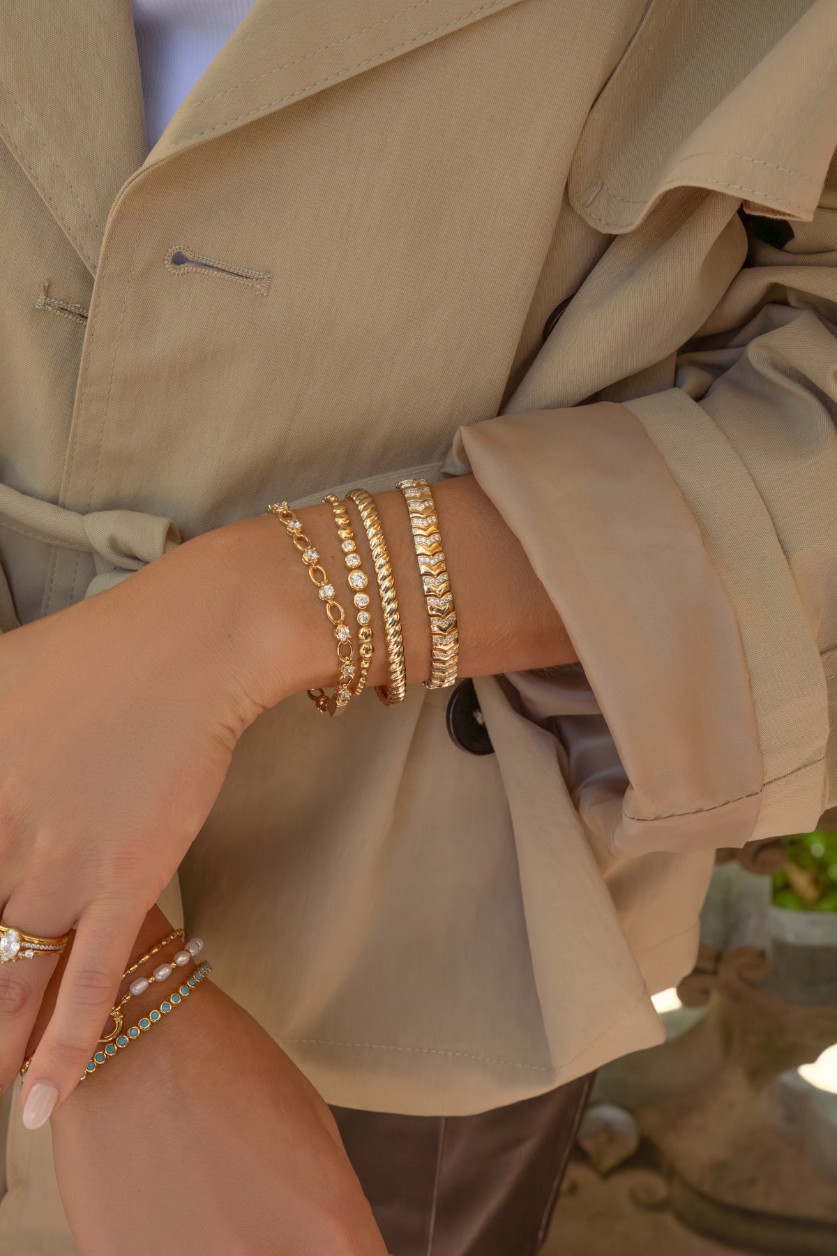 A close-up of a person wearing multiple gold bracelets on one wrist, including a tennis bracelet, chain bracelet, and textured link bracelet, styled with a beige trench coat.
