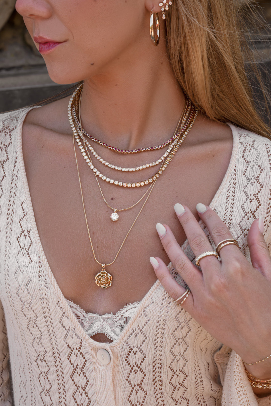 A close-up lifestyle shot of a woman wearing layered gold necklaces, including a rose pendant, beaded chains, and a pearl charm, along with gold rings and hoop earrings.